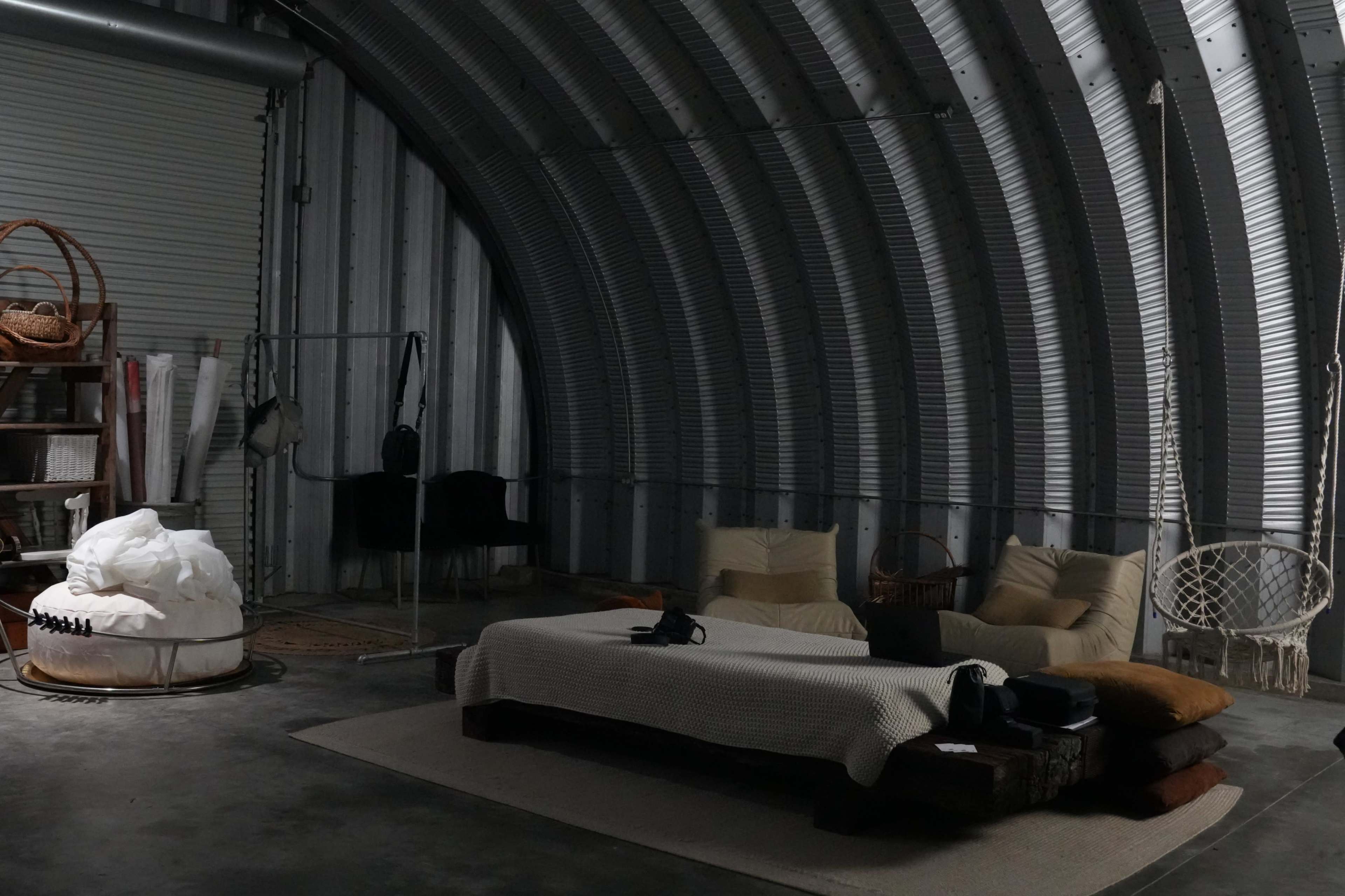 The image shows a spacious, industrial-style living area with curved metal walls, featuring a low-profile couch, a hanging chair, and various textured pillows arranged on a rug.