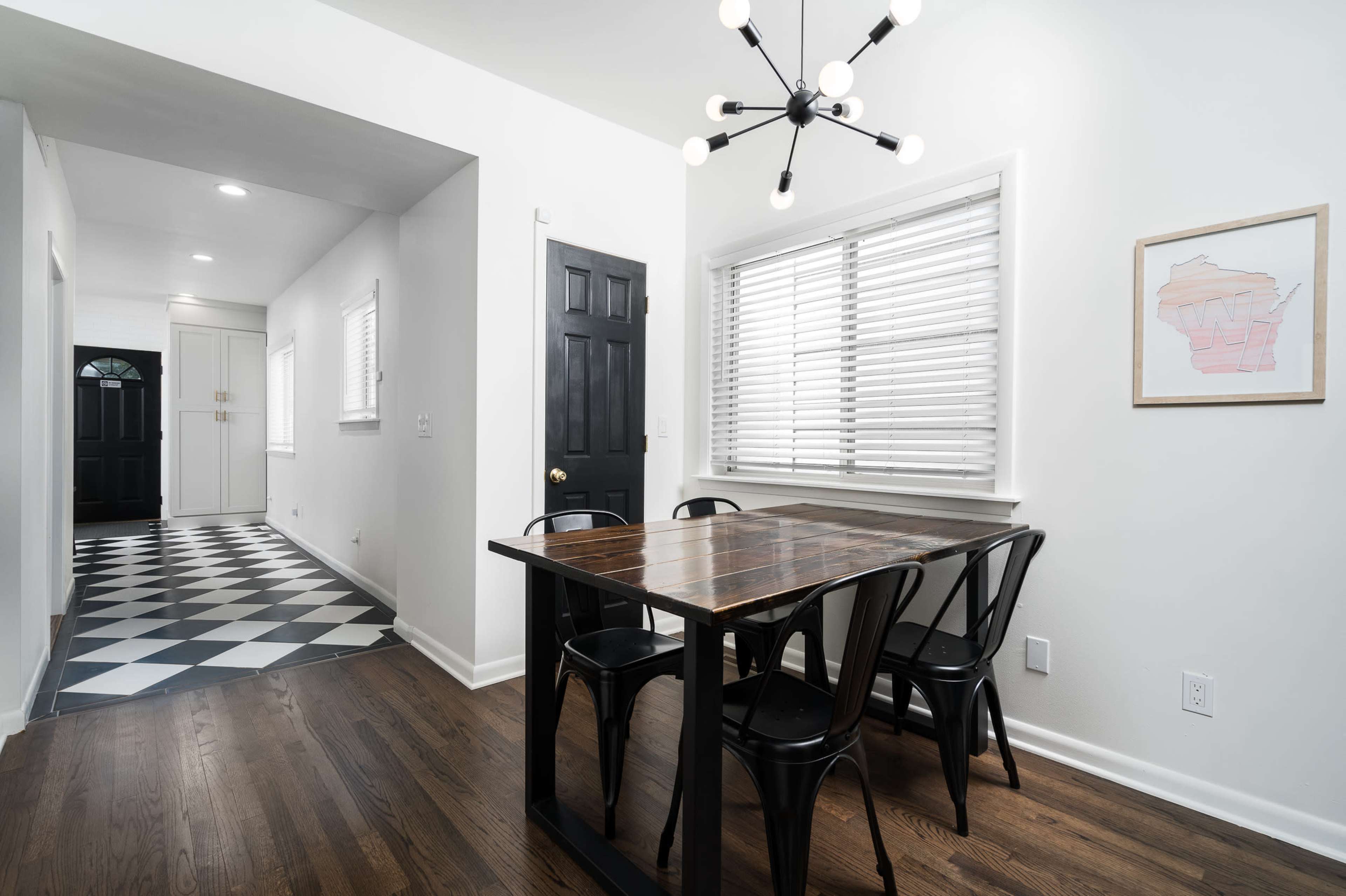 A modern dining area features a wooden table with black chairs, adjacent to a hallway with black-and-white checkered flooring and a doorway leading outside.