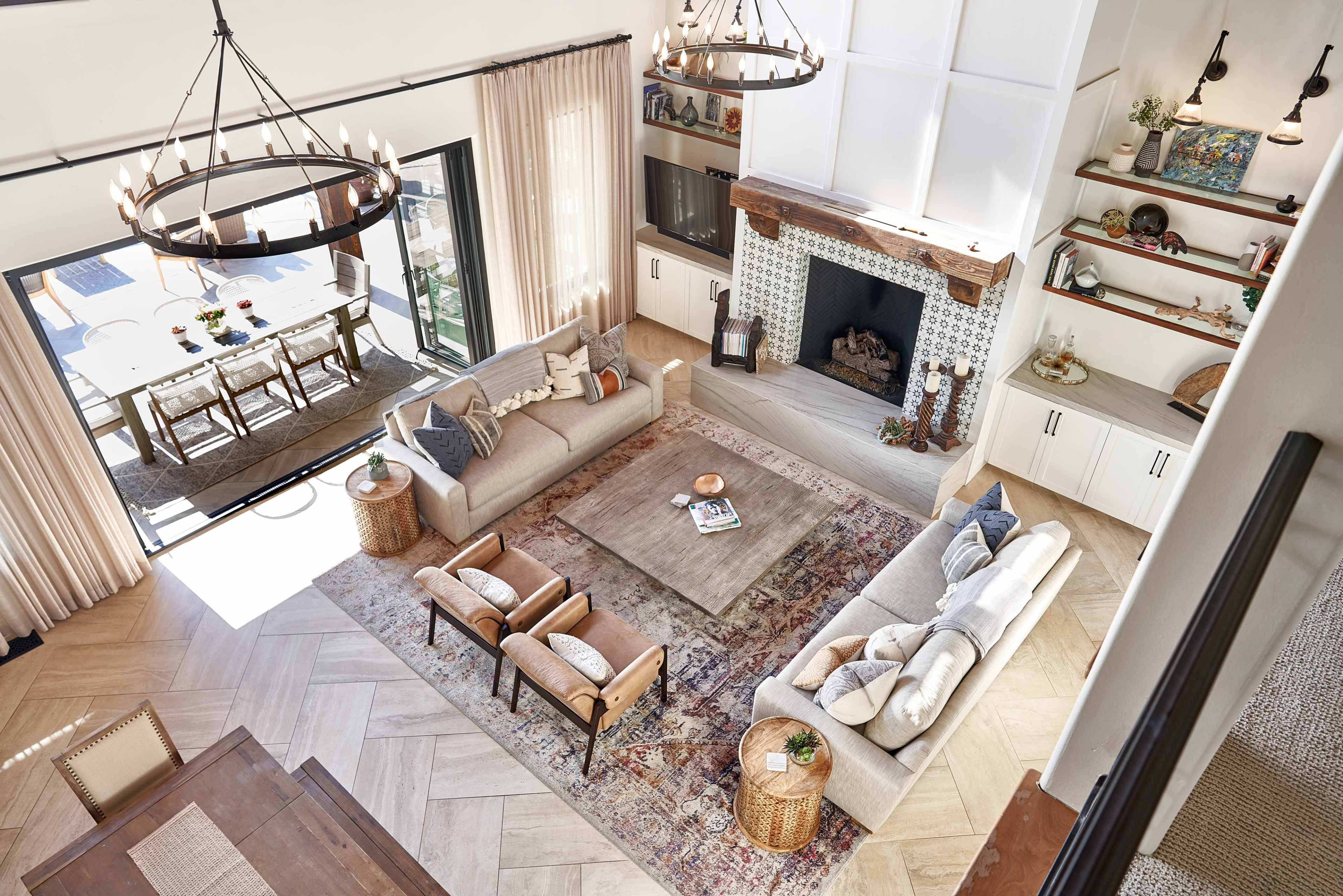 The image features a spacious living room with two sofas, armchairs, a coffee table, and a large area rug, all arranged around a central fireplace, with large windows displaying a dining area in the background.