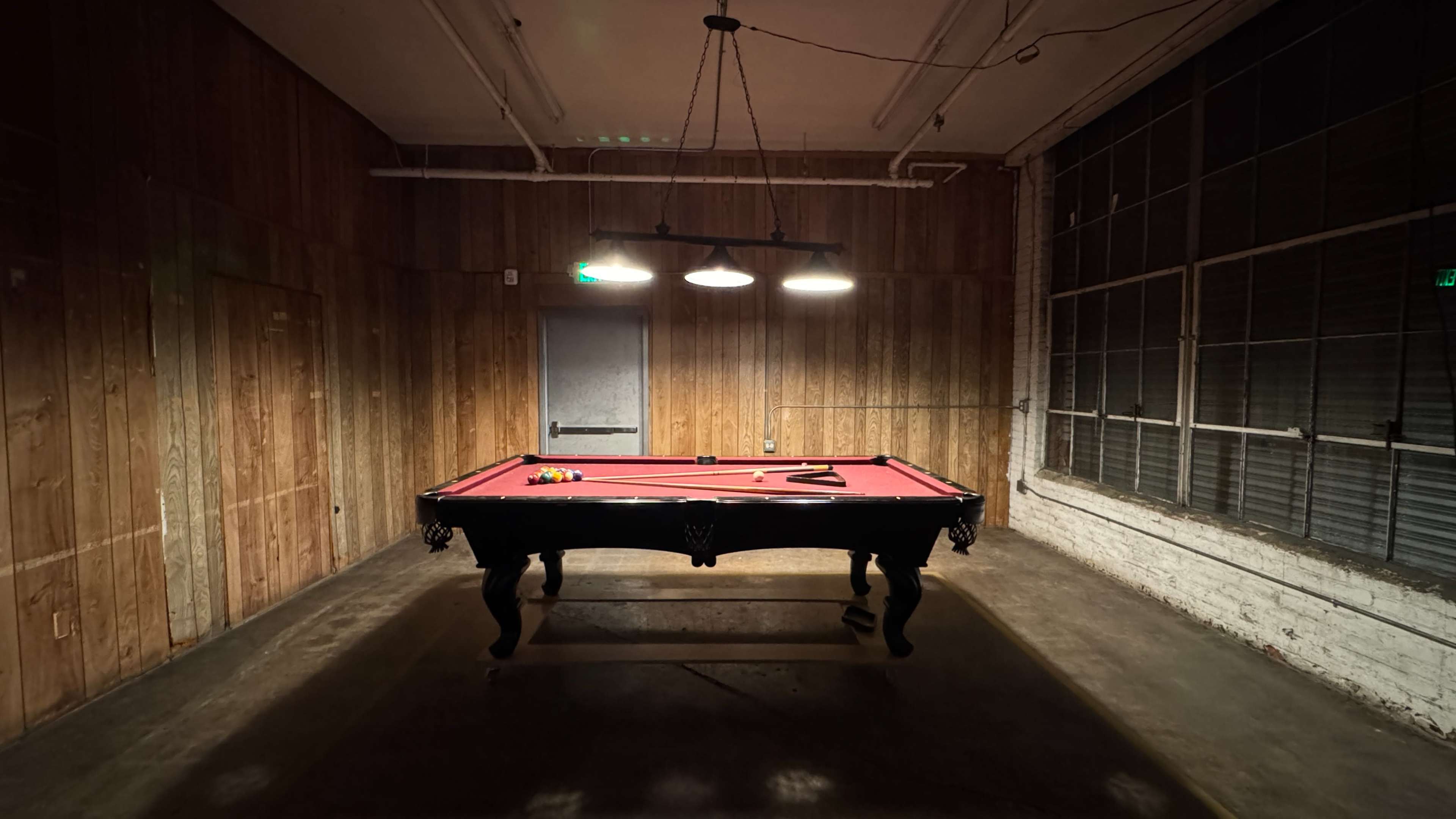 A pool table with a red felt surface is illuminated by overhead lights in a dimly lit, wood-paneled room with a large window.