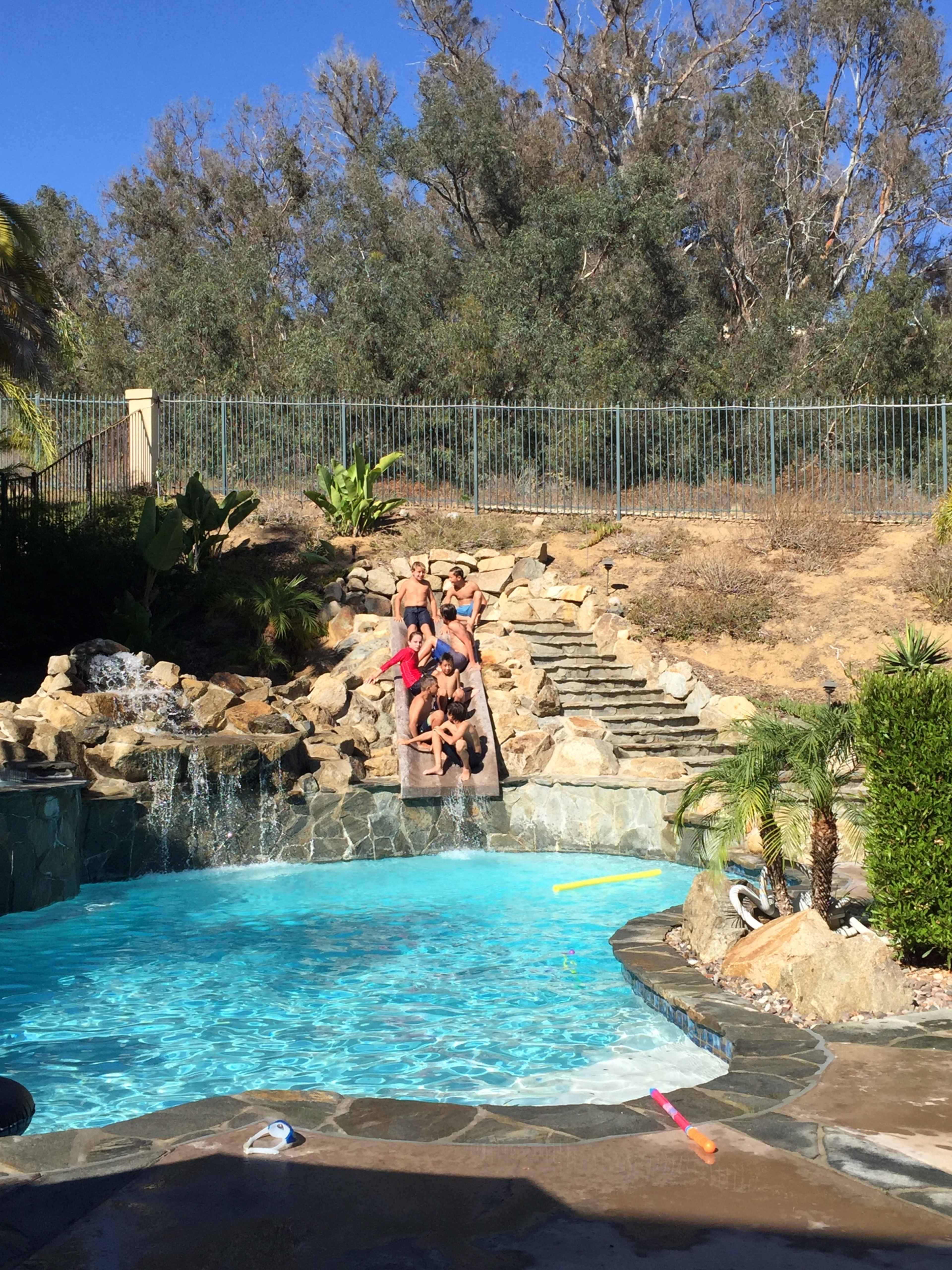 A group of children sits on a stone ledge beside a swimming pool surrounded by greenery and rocks.