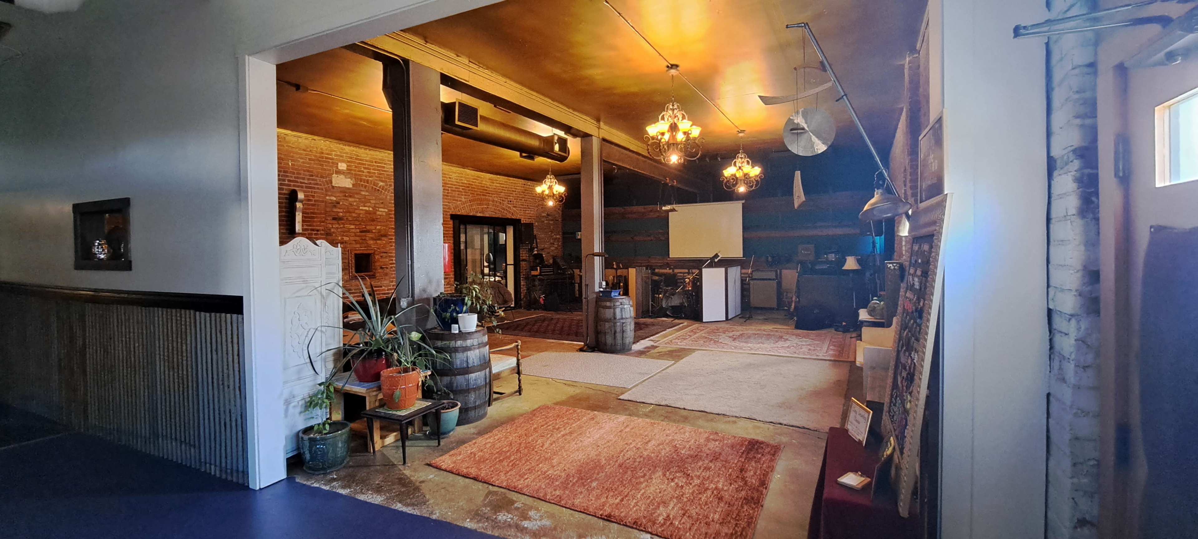An interior space with brick walls, wooden beams, chandeliers, and various rugs, set up for events with plants and barrels as decor.
