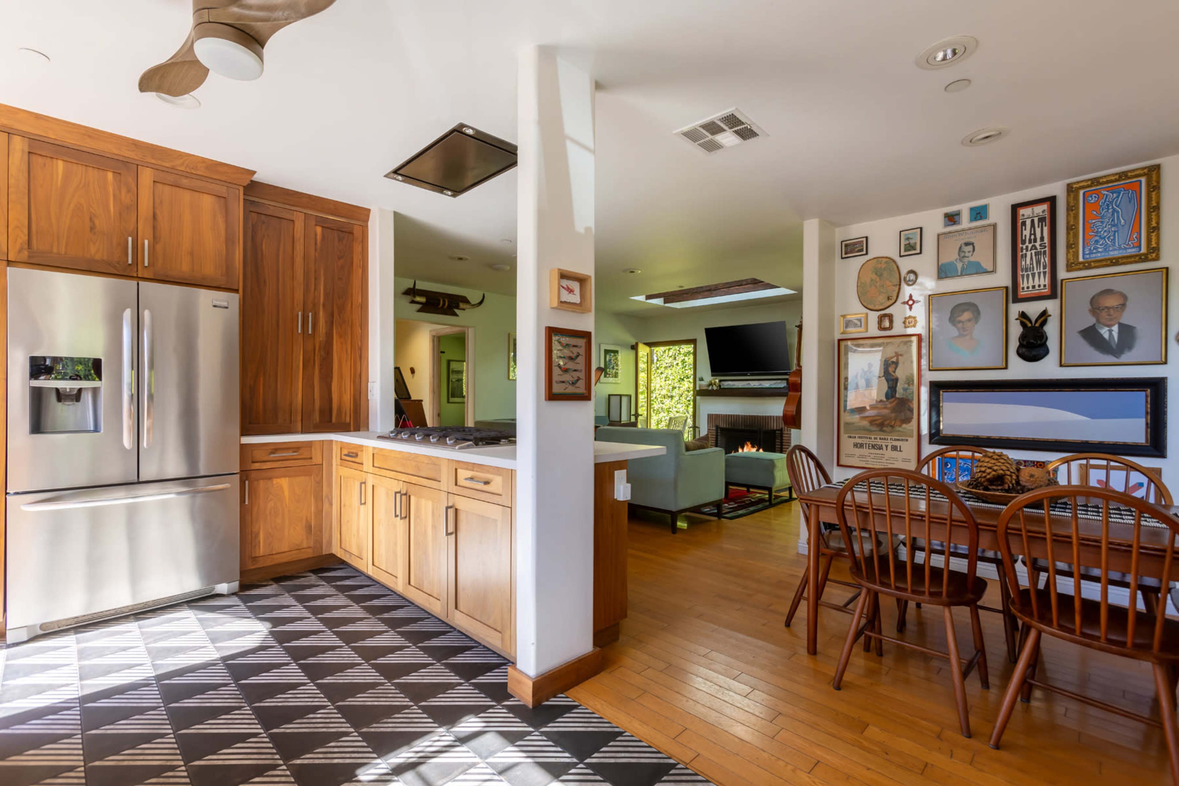 The image shows a modern kitchen with wooden cabinetry, stainless steel appliances, and a dining area adorned with various framed artworks and a television.