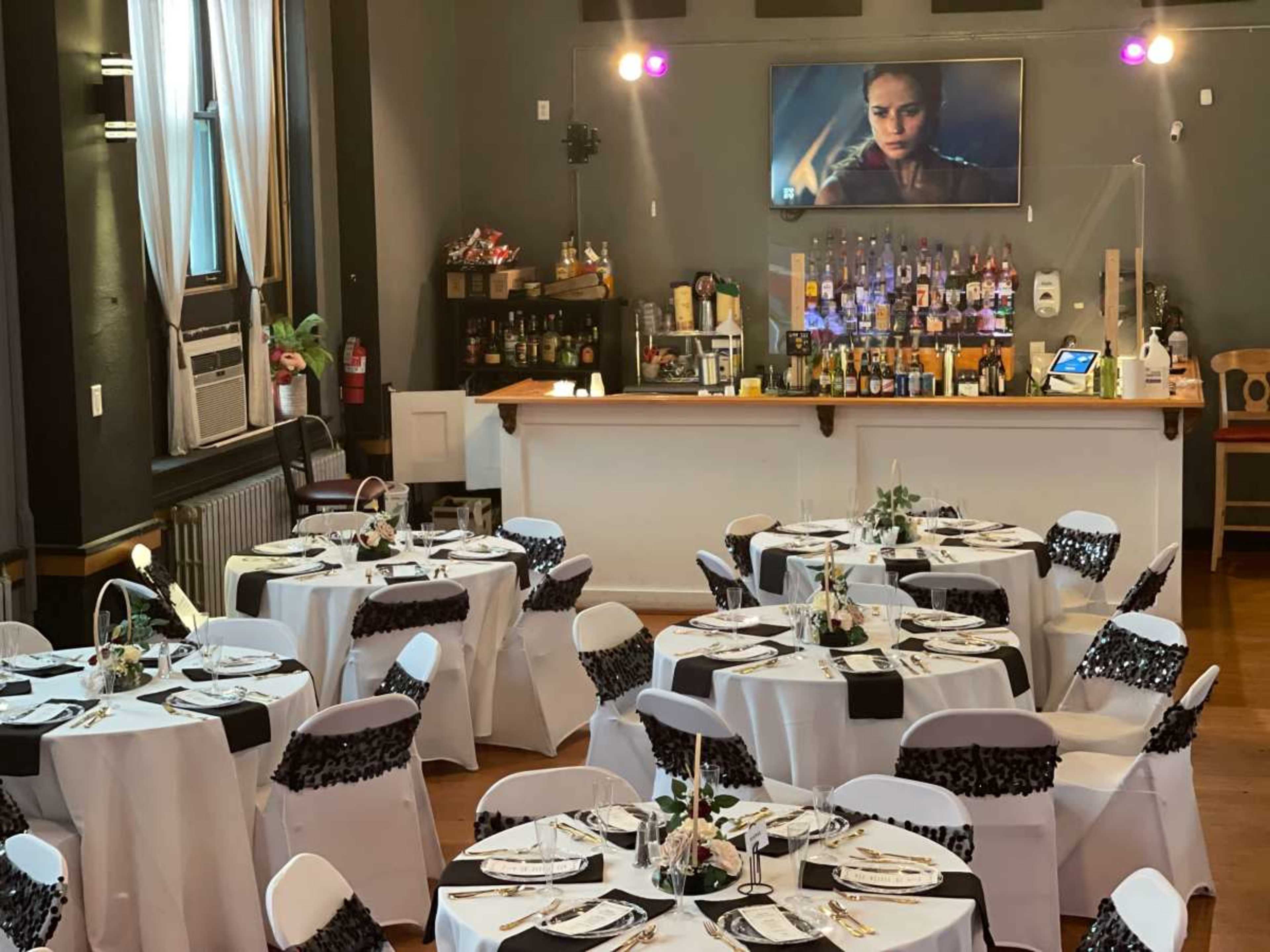 The image shows a dining area with elegantly set tables, each decorated with white tablecloths and black accents, and a bar in the background displaying various beverages.