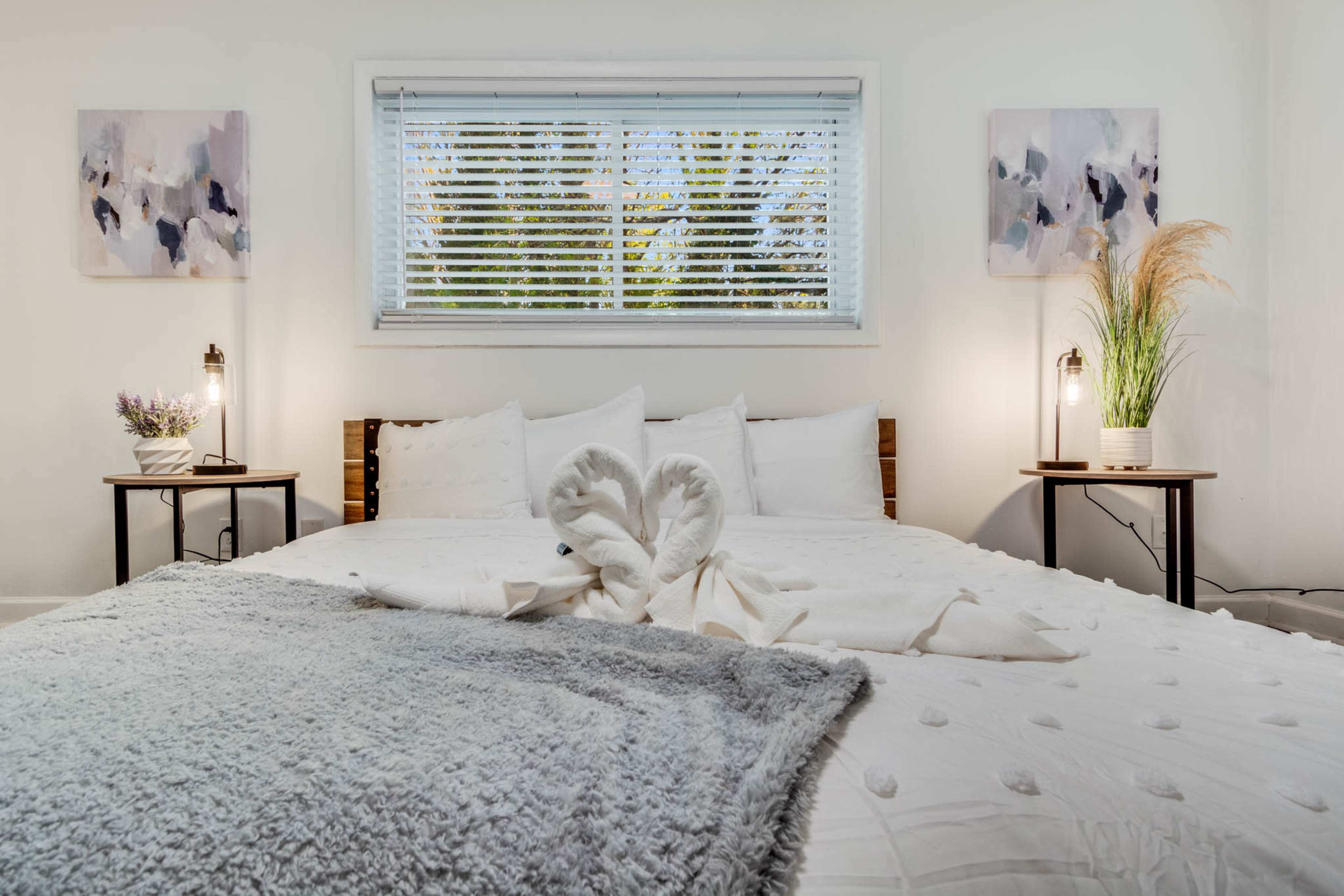 A neatly made bed with decorative pillows is positioned in a bright bedroom featuring two paintings on the walls and potted plants on bedside tables.