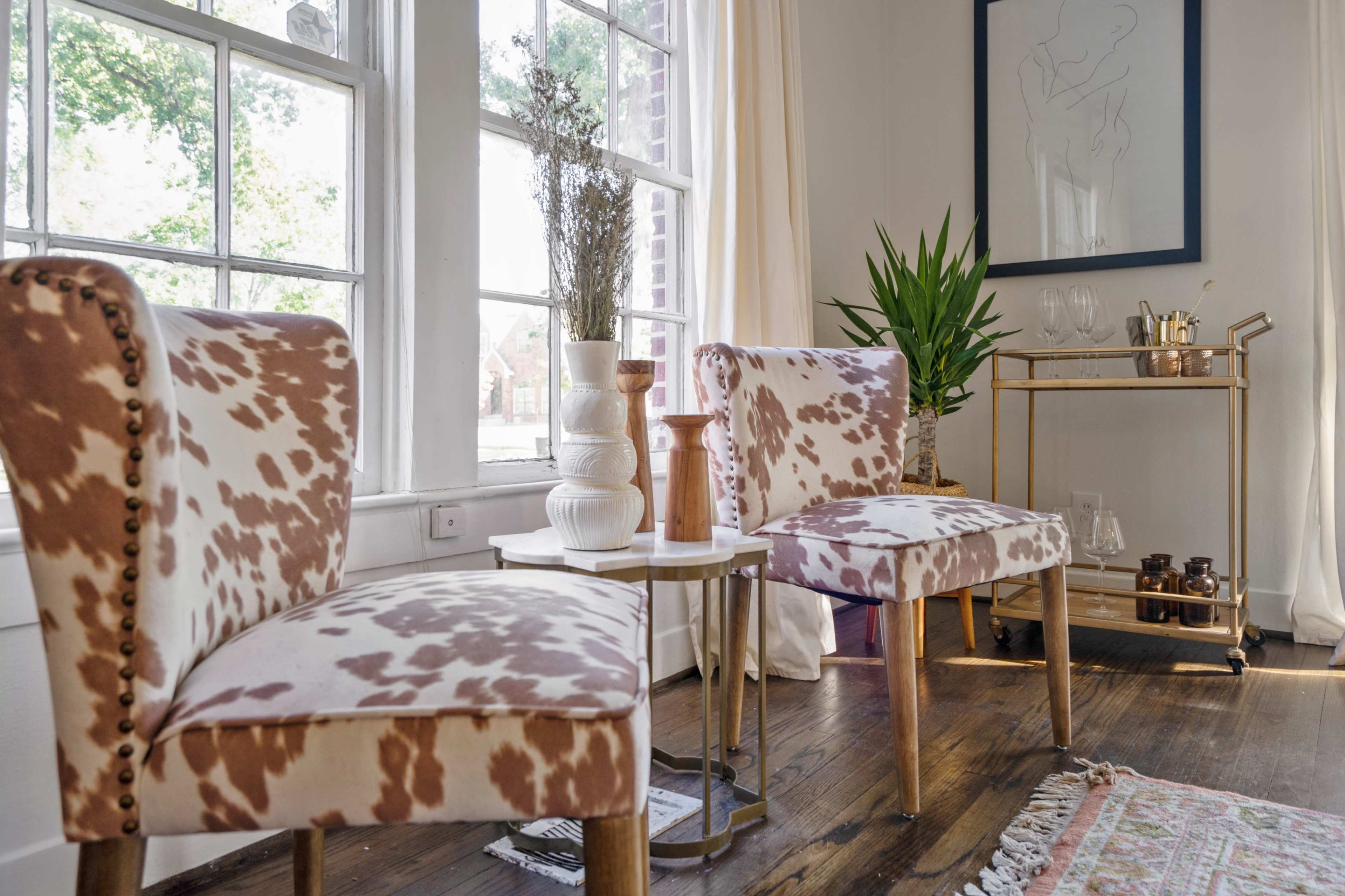 Two patterned chairs set beside a small table near large windows, with a potted plant and a bar cart in the background.