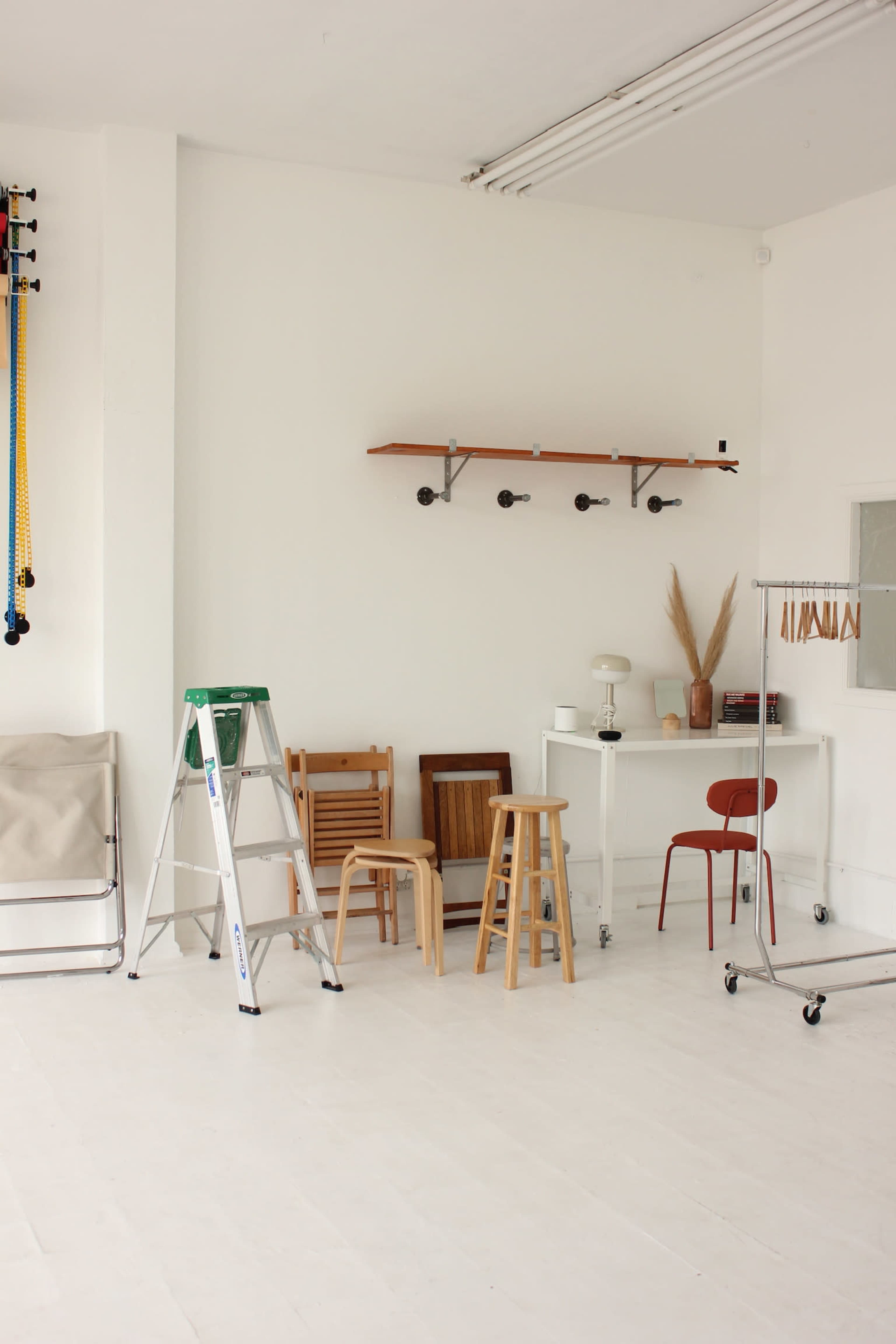 The image shows a minimalistic interior space featuring a ladder, various wooden chairs, a small table, and a shelf mounted on a white wall.