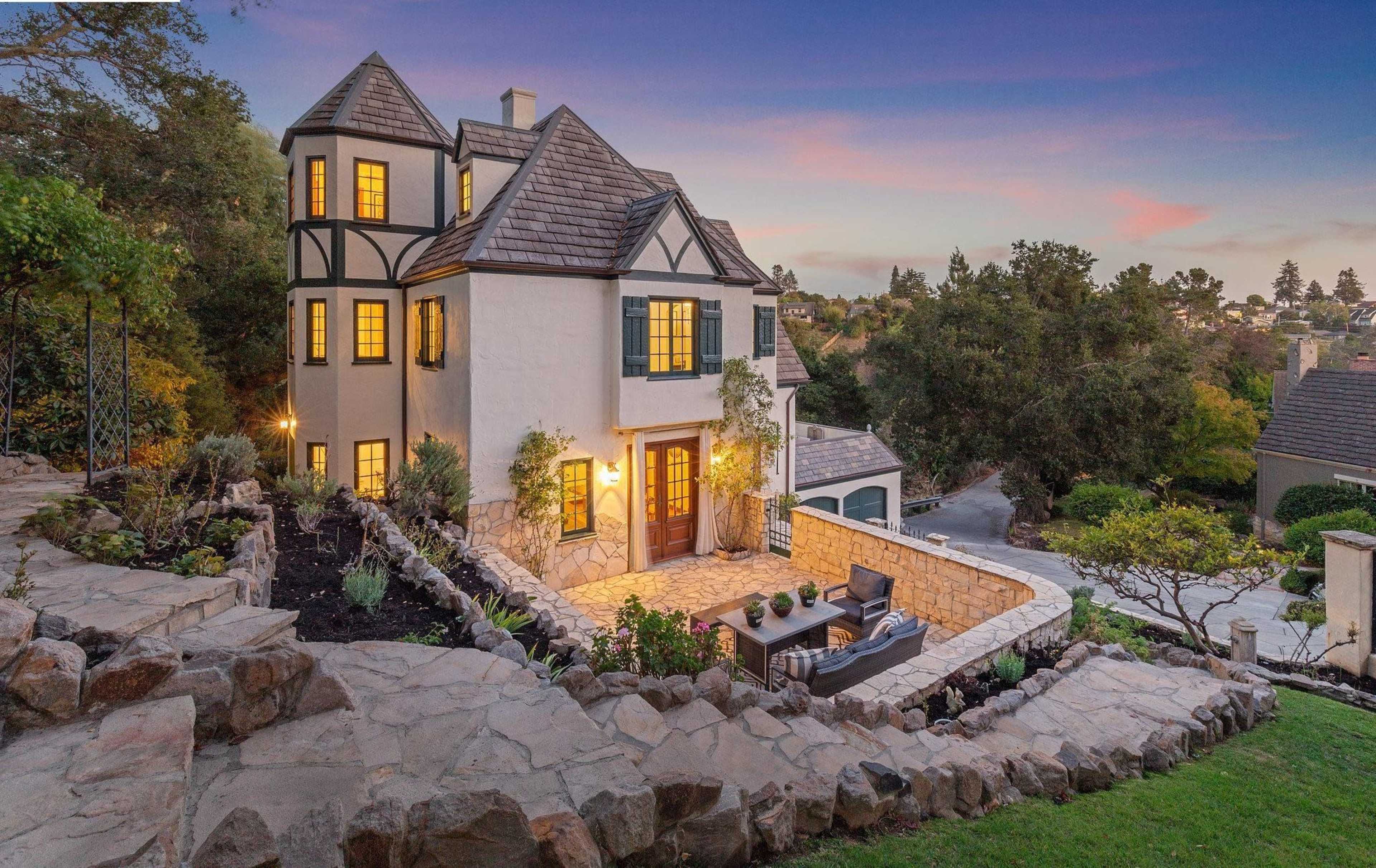 A two-story house with a mix of stone and stucco features a turret, surrounded by landscaped gardens and a stone pathway.