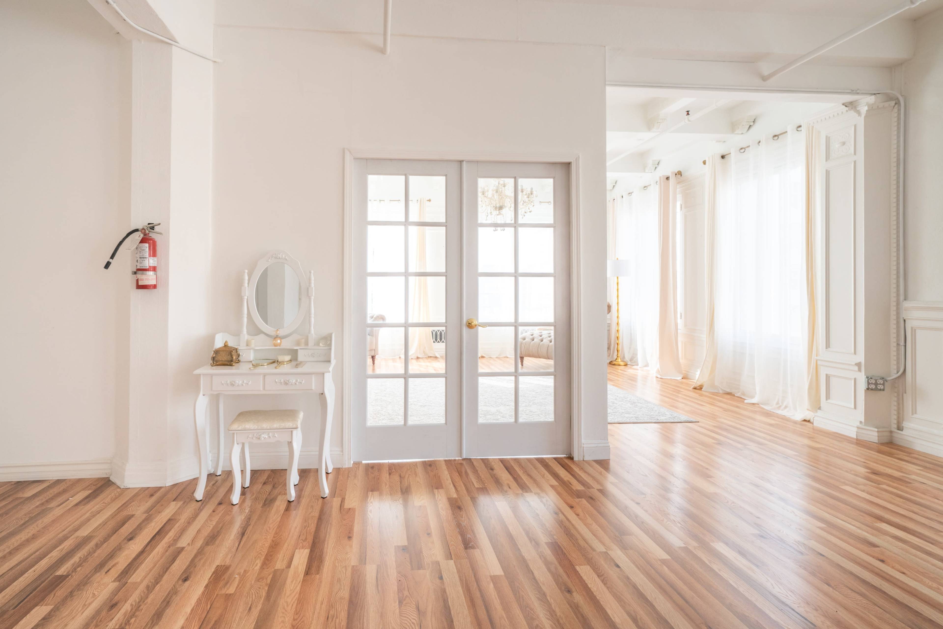 A bright, spacious room with wooden flooring, a white dresser with a mirror, and large windows draped in sheer curtains.