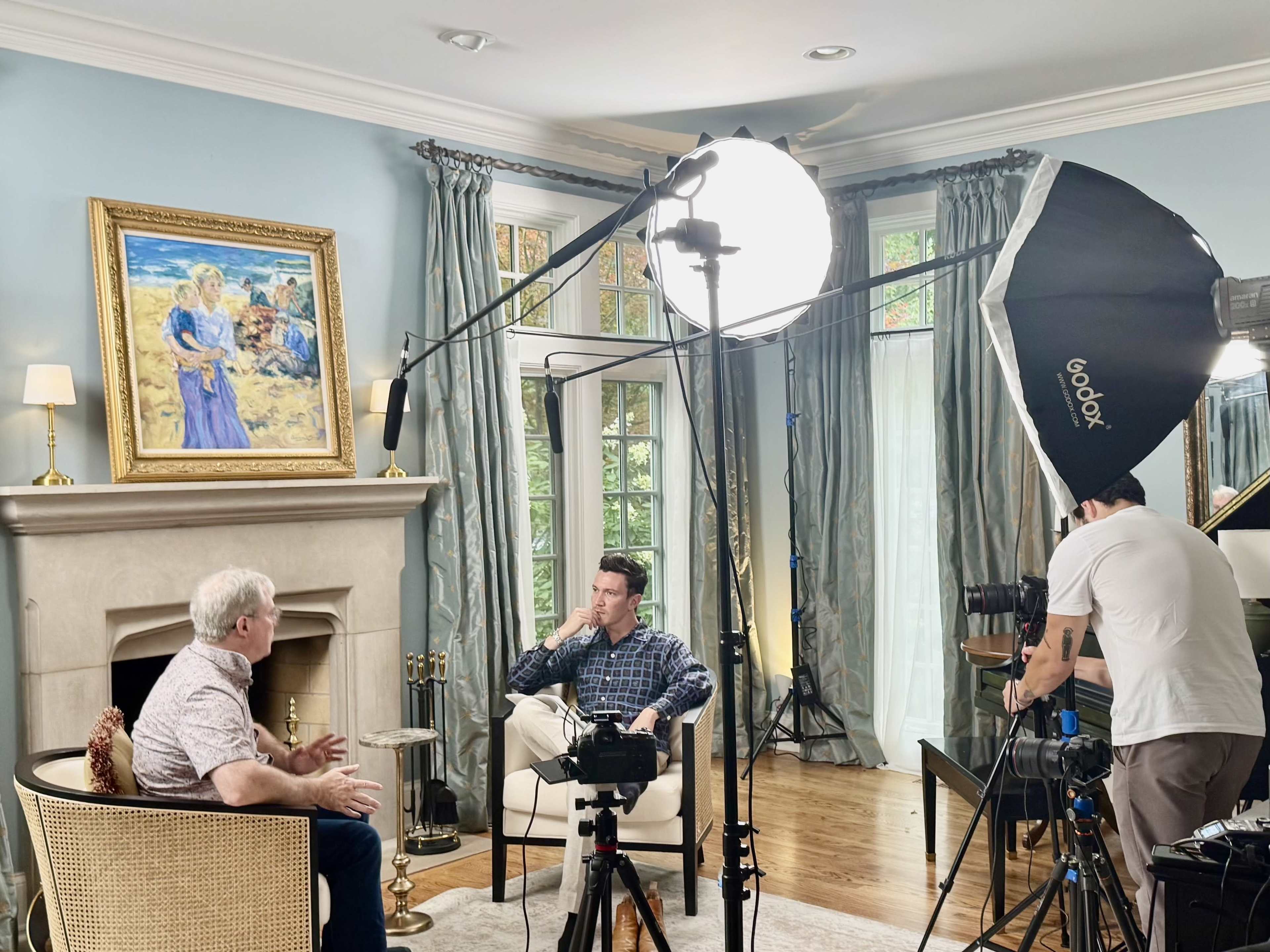 A man is being interviewed in a well-lit living room, with a camera and lighting equipment set up around him.