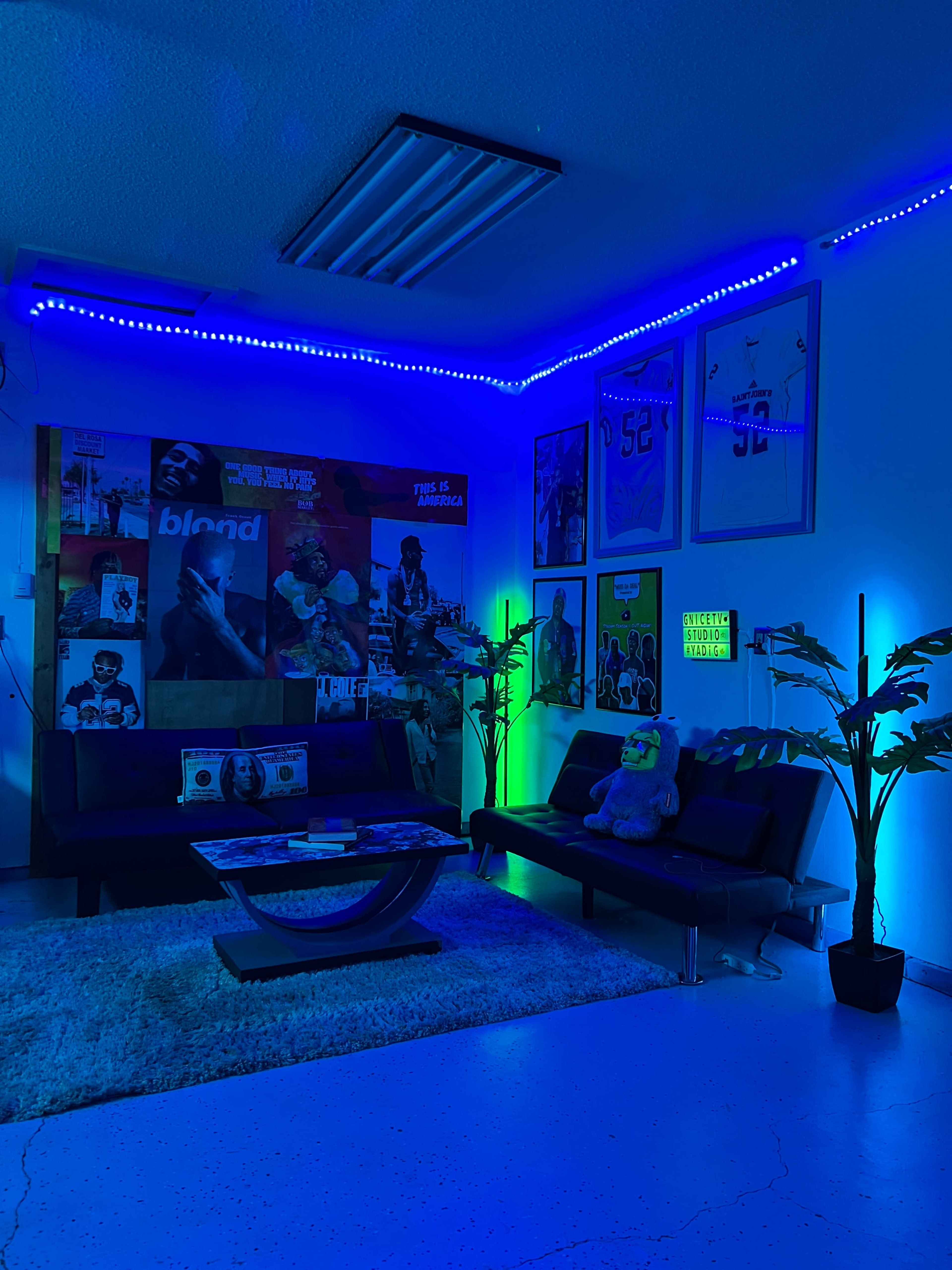 The image shows a cozy lounge area with a dark sofa, colorful posters on the walls, and blue ambient lighting.