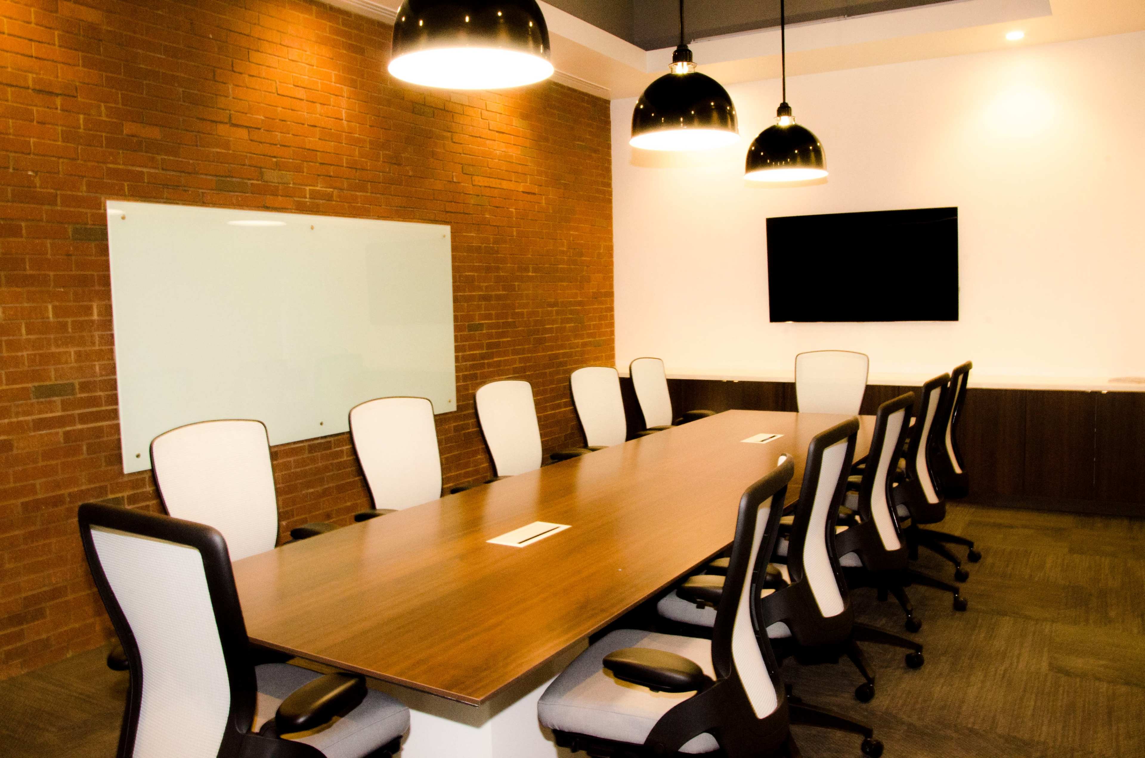 A modern conference room features a long wooden table surrounded by white and black ergonomic chairs, with a whiteboard and a screen mounted on the wall.