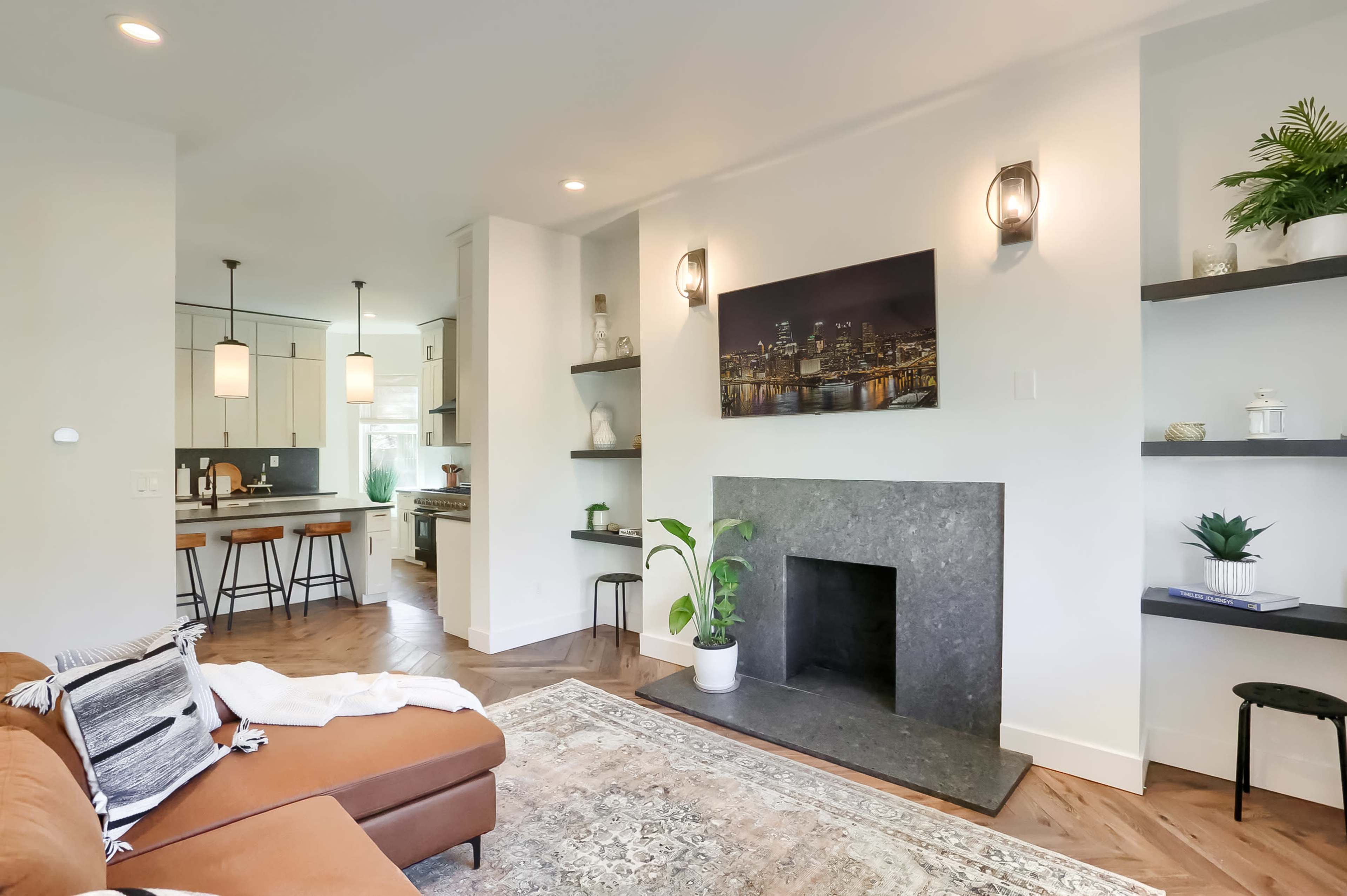 A modern living room with a brown sofa, a black stone fireplace, and open shelves, connected to a kitchen area with pendant lighting.