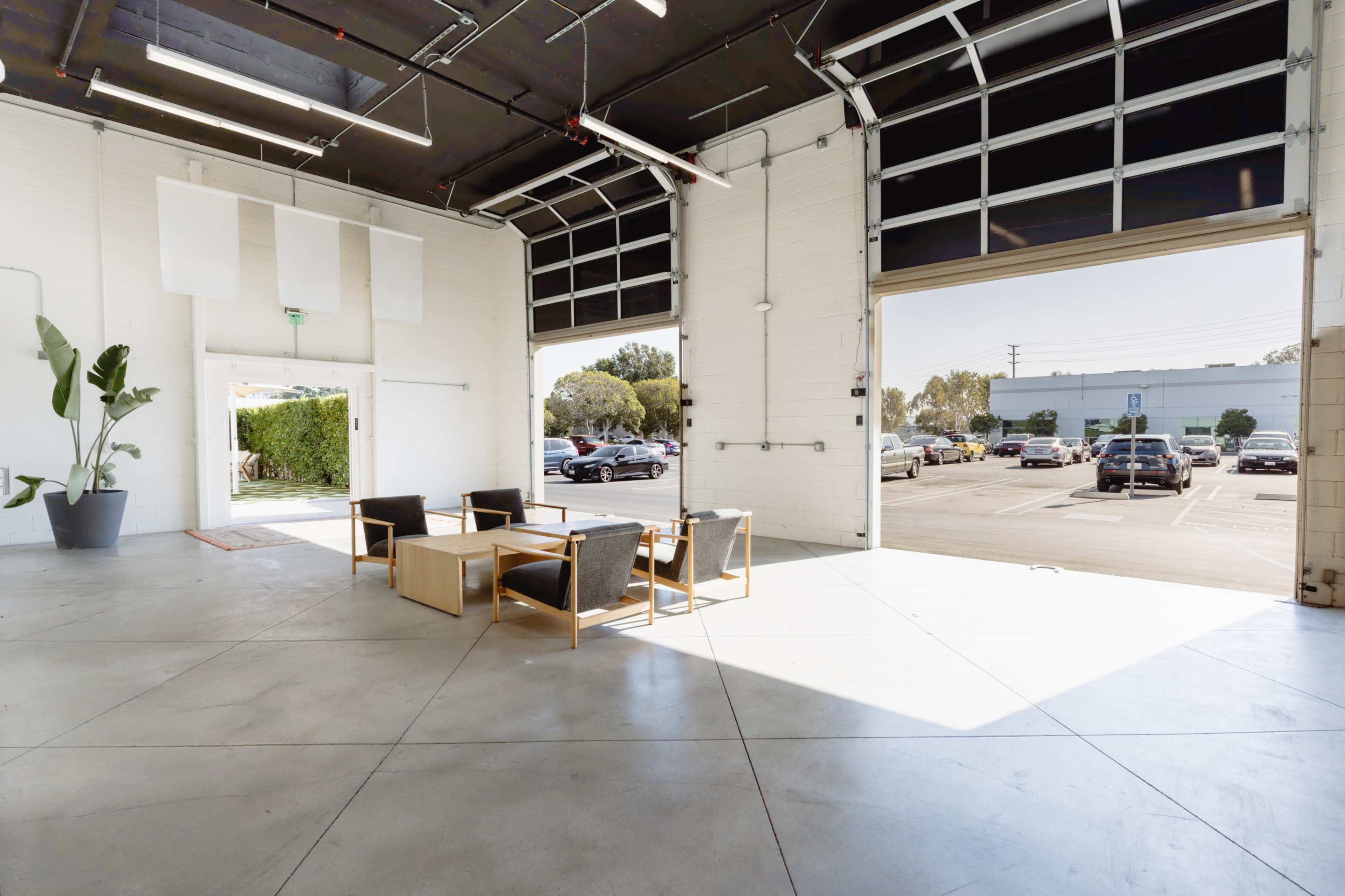 The image shows an empty garage-like space with large doors, a seating area with chairs and a table, and cars visible outside through the open entrance.