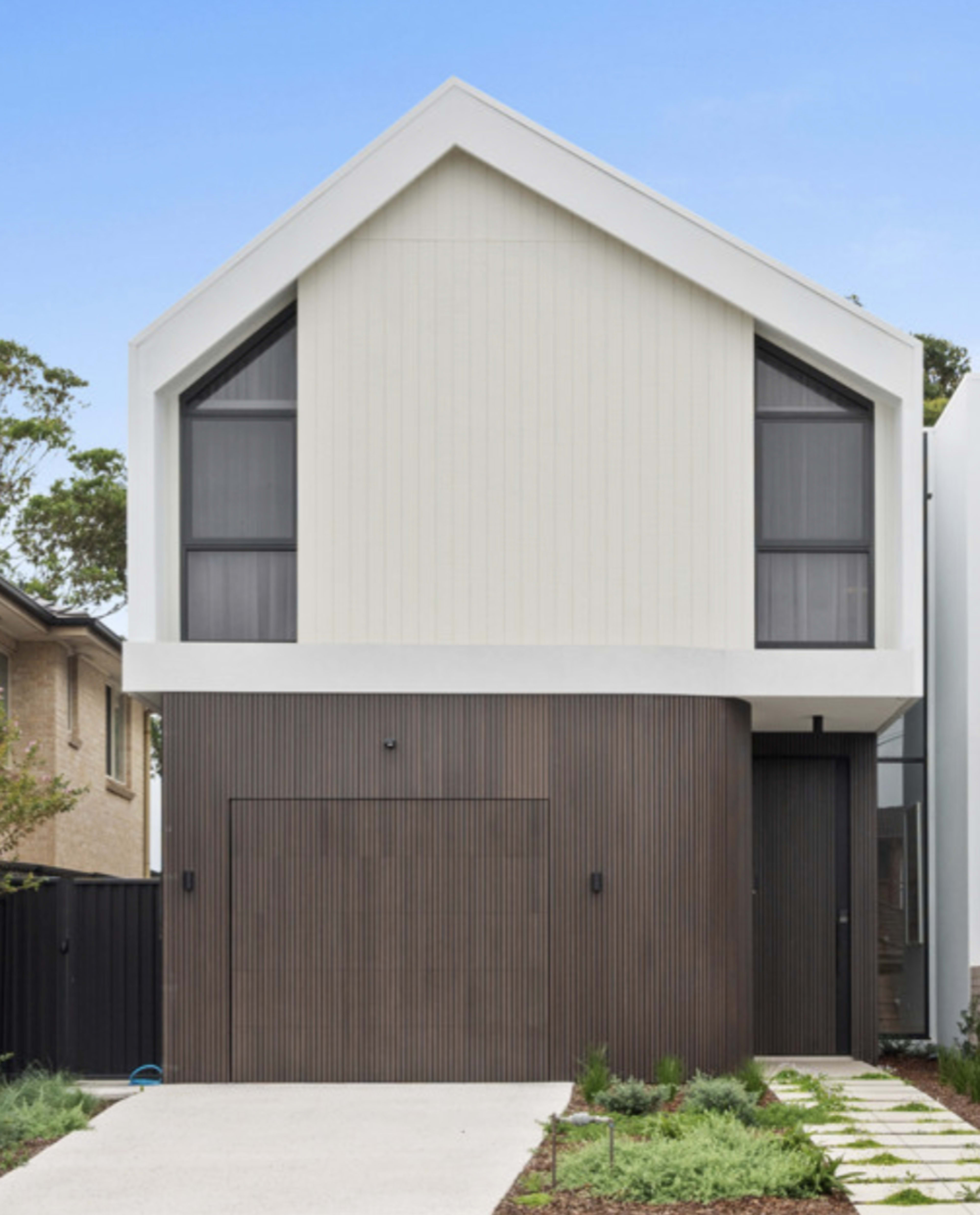 The image shows a modern two-story house with a sloped roof, featuring a combination of white and brown materials, and a landscaped front yard.