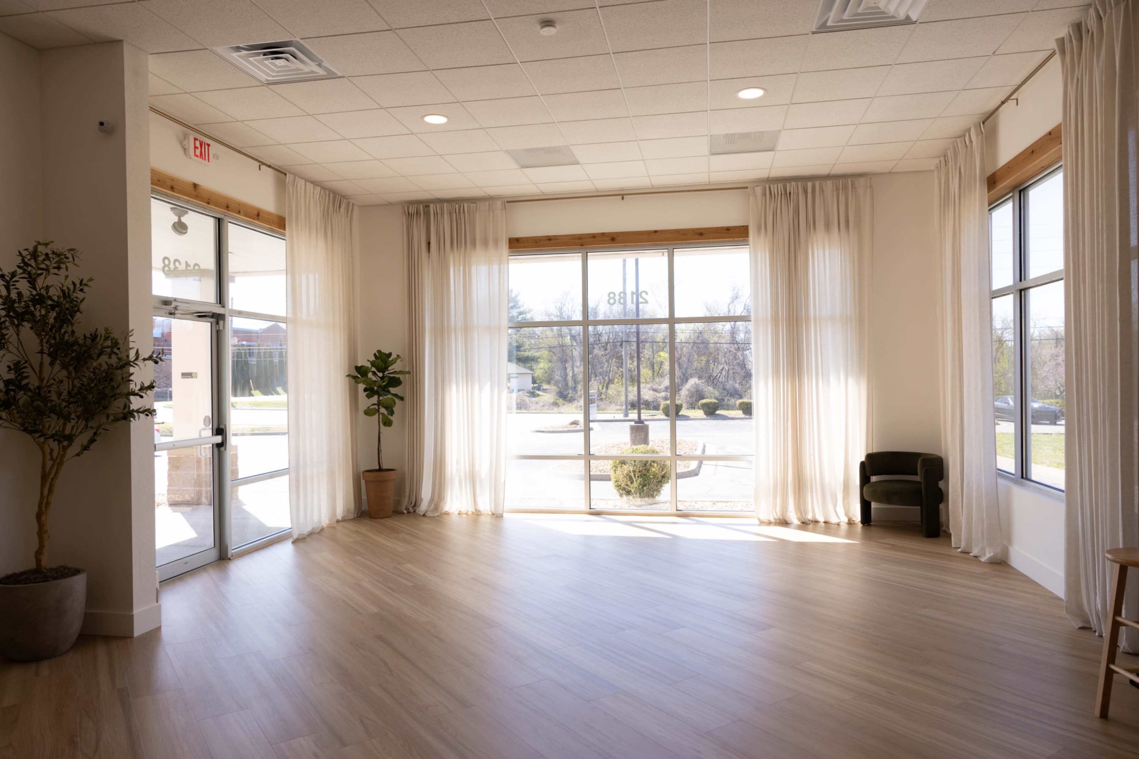 A bright, empty room with large windows, white curtains, and wooden flooring, featuring a small plant and a chair near the entrance.