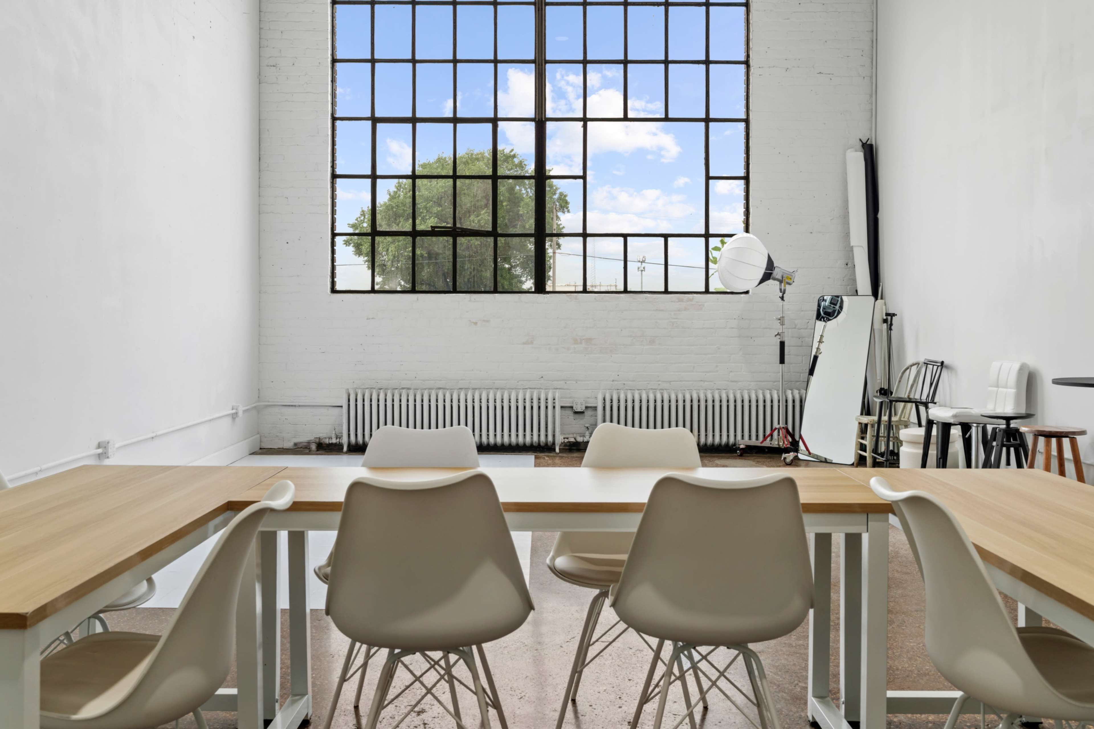 The image shows a spacious room with large windows, a wooden table surrounded by white chairs, and a visible radiator along the wall.
