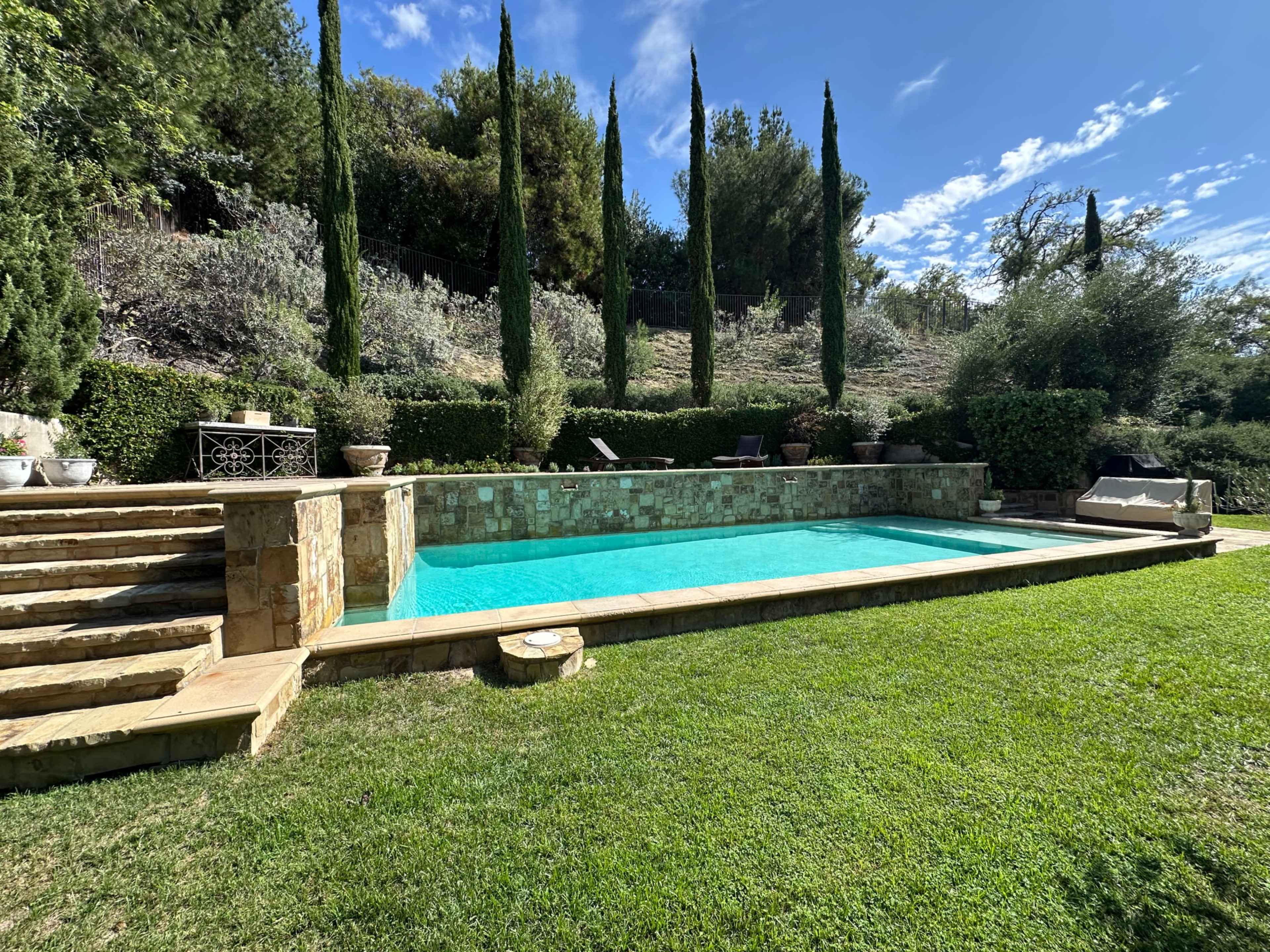 A rectangular swimming pool surrounded by green grass and tall trees, with stone steps leading down to the water.