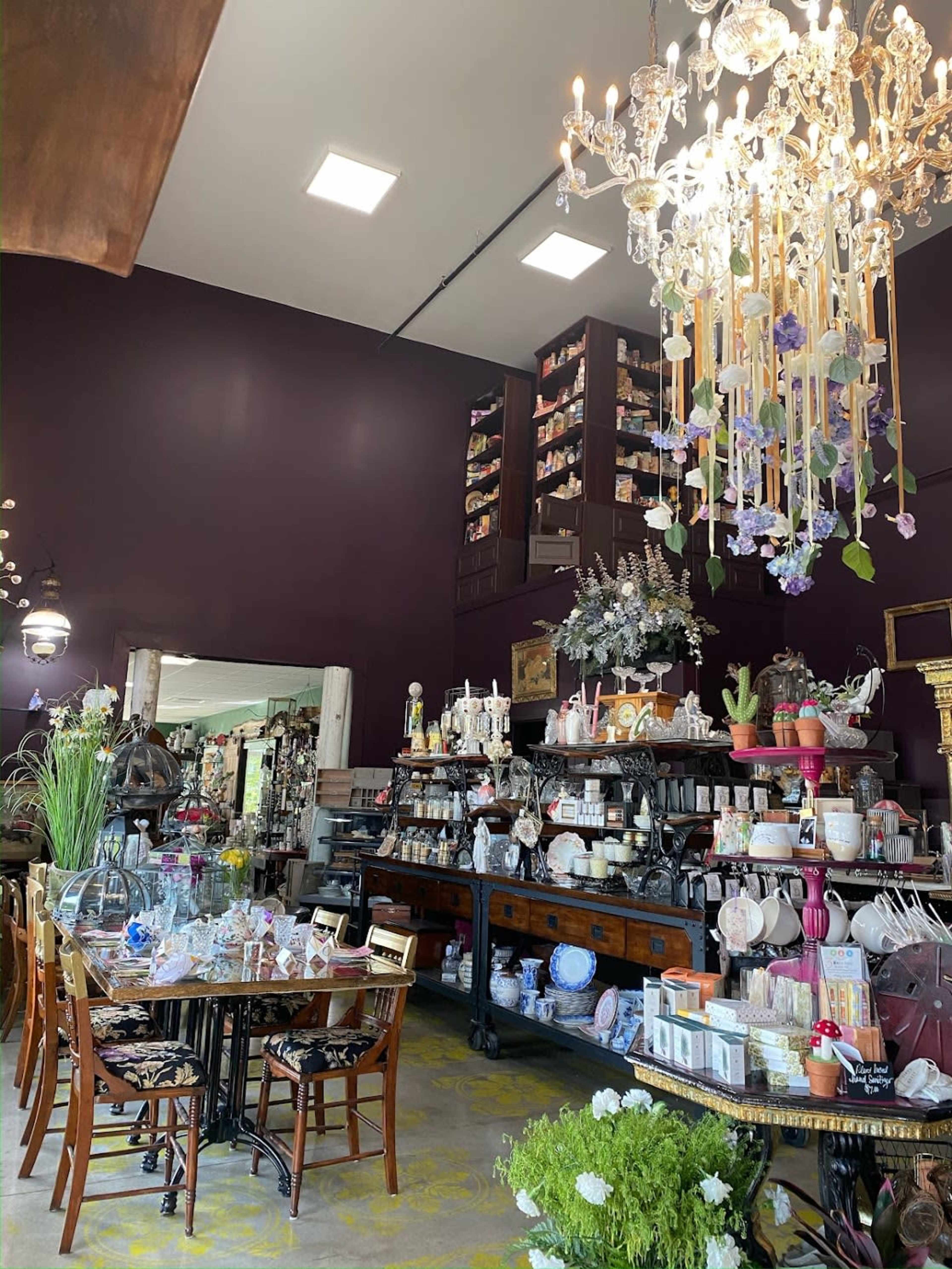 An elegantly arranged vintage shop featuring a chandelier, displays of various decorative items, and a dining table set with glassware and dishware.