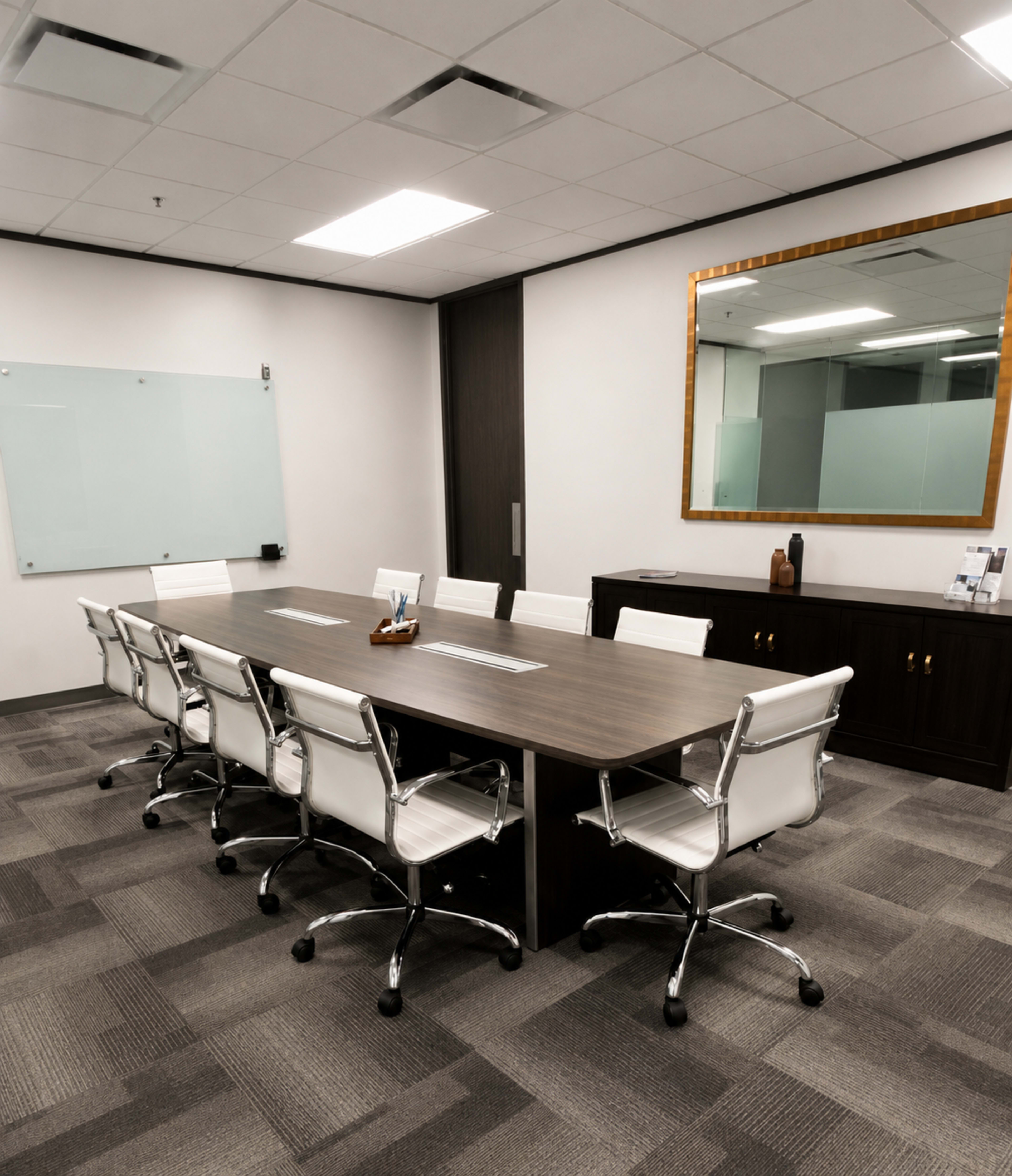 The image shows a modern conference room with a long wooden table surrounded by white rolling chairs, a large mirror on one wall, and a glass whiteboard.