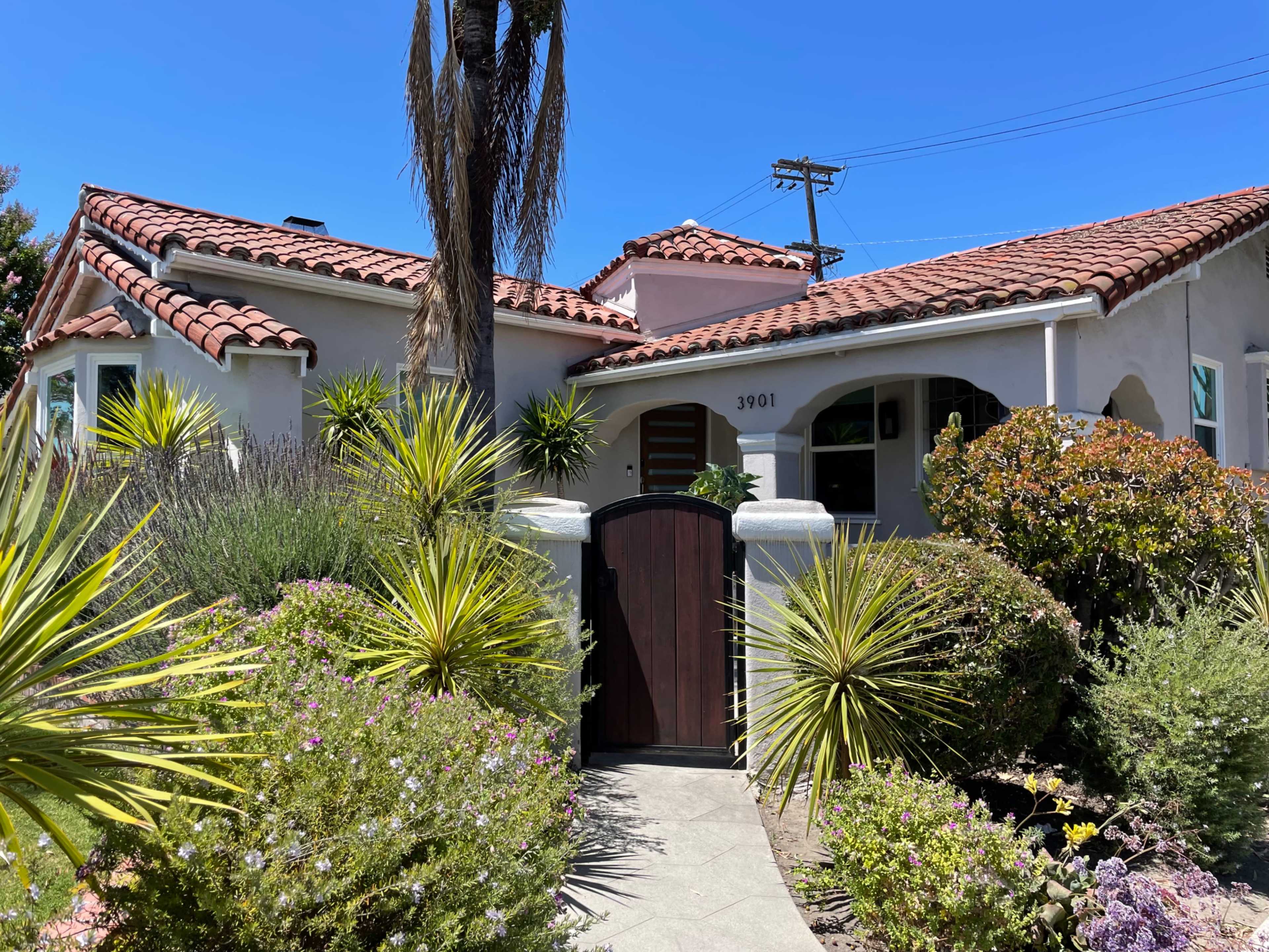 A Mediterranean-style house with a red-tiled roof, surrounded by various plants and a wooden gate.
