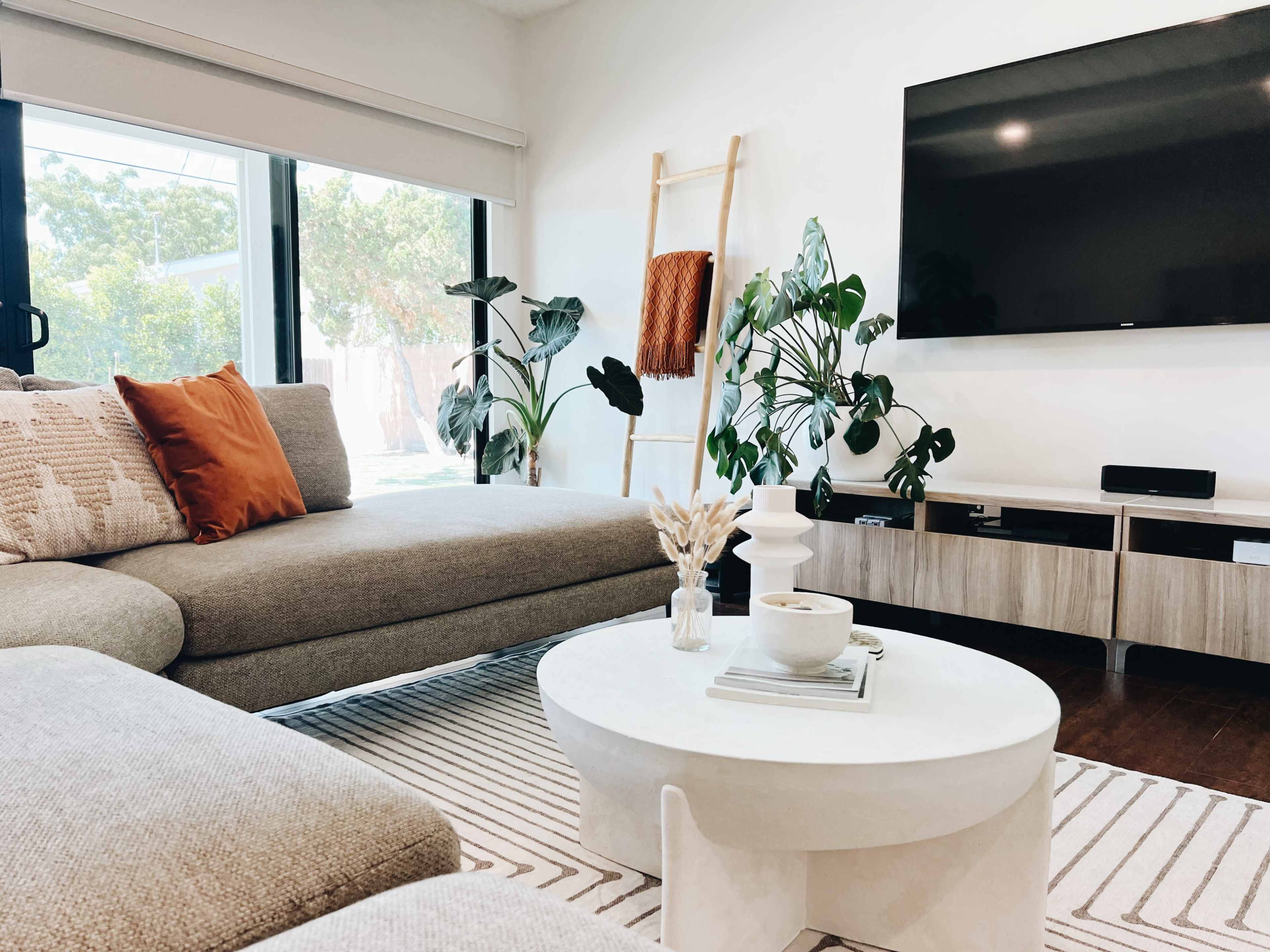 A modern living room with a sectional sofa, a round coffee table, and a large TV mounted on the wall, surrounded by indoor plants and natural light.