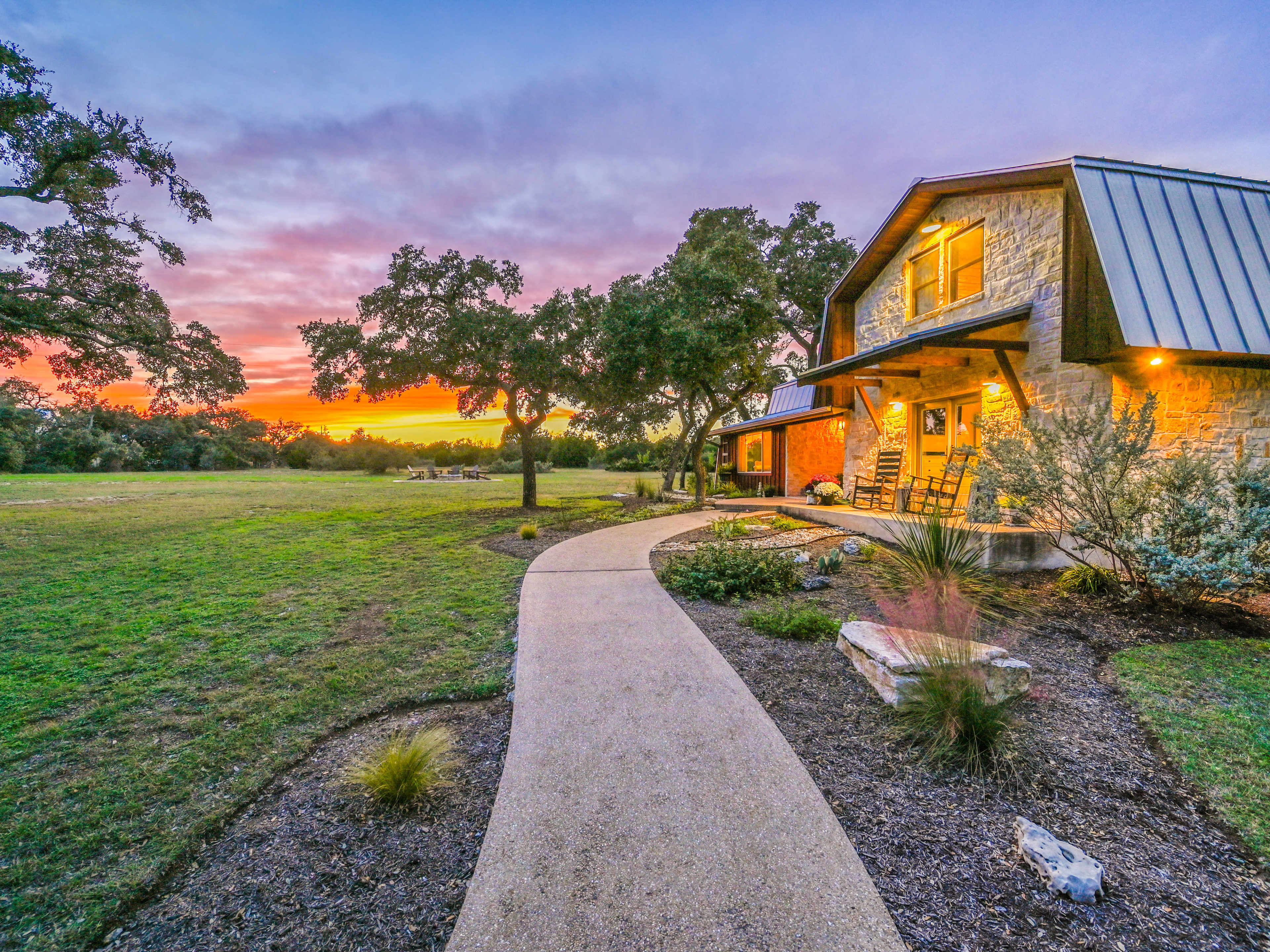 A curved path leads to a stone house with a metal roof, surrounded by trees and a colorful sunset in the background.