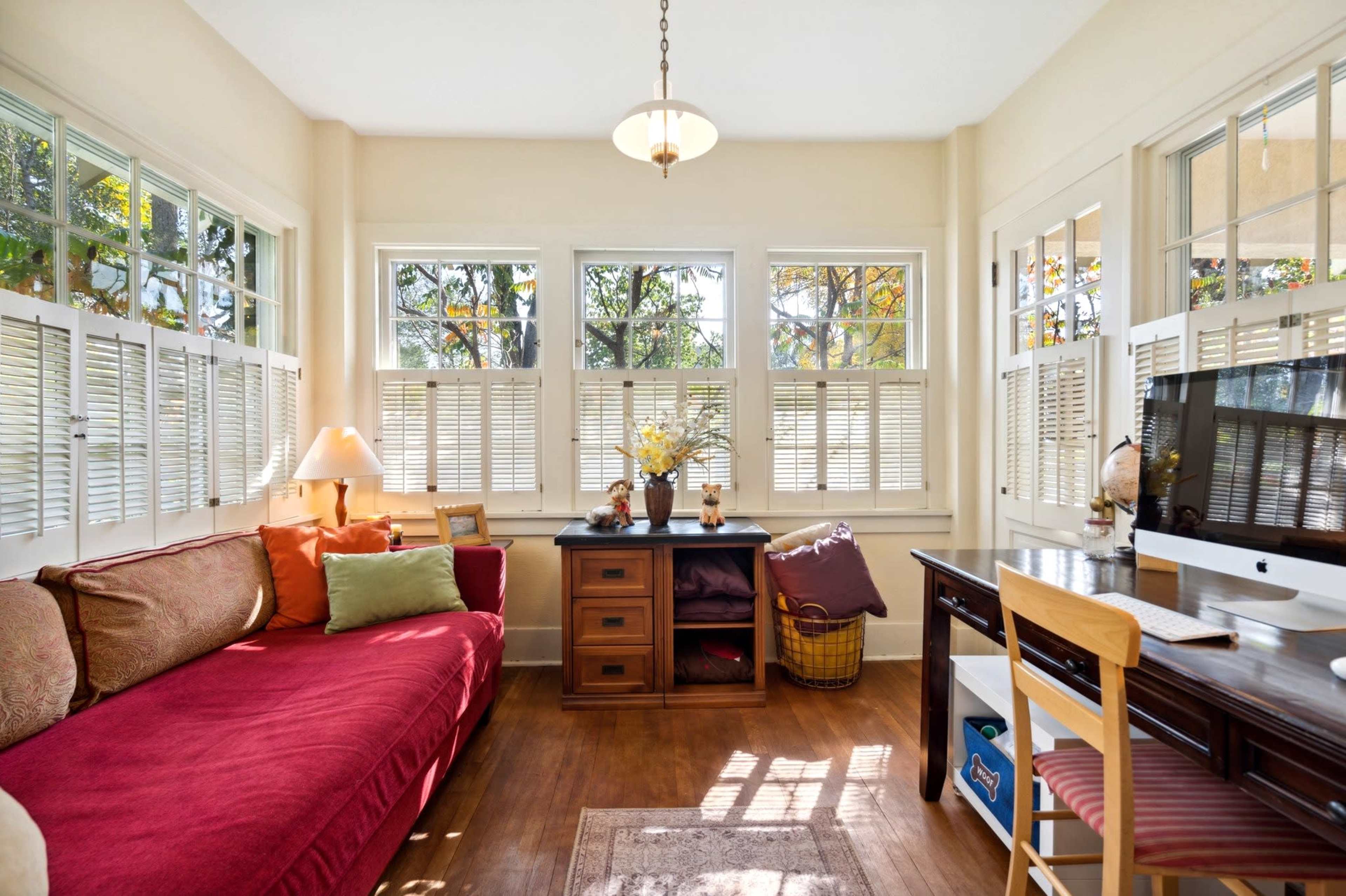 The image shows a sunlit room with large windows, a red couch adorned with cushions, a wooden desk with a computer, and a decorative table with arranged flowers.