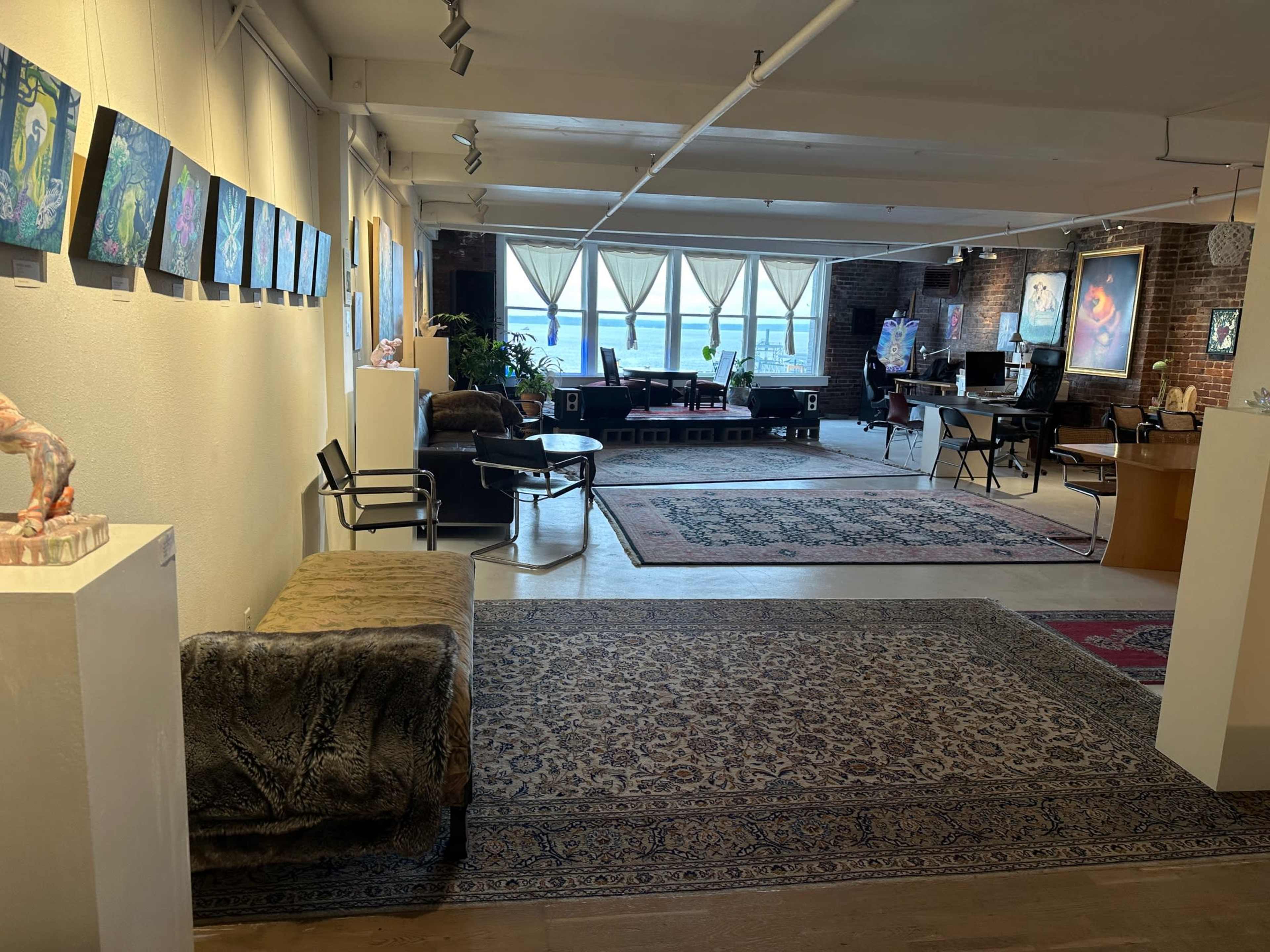 An art gallery or studio space featuring colorful paintings on the walls, a seating area with a plush sofa, and various workstations in a well-lit, airy environment.