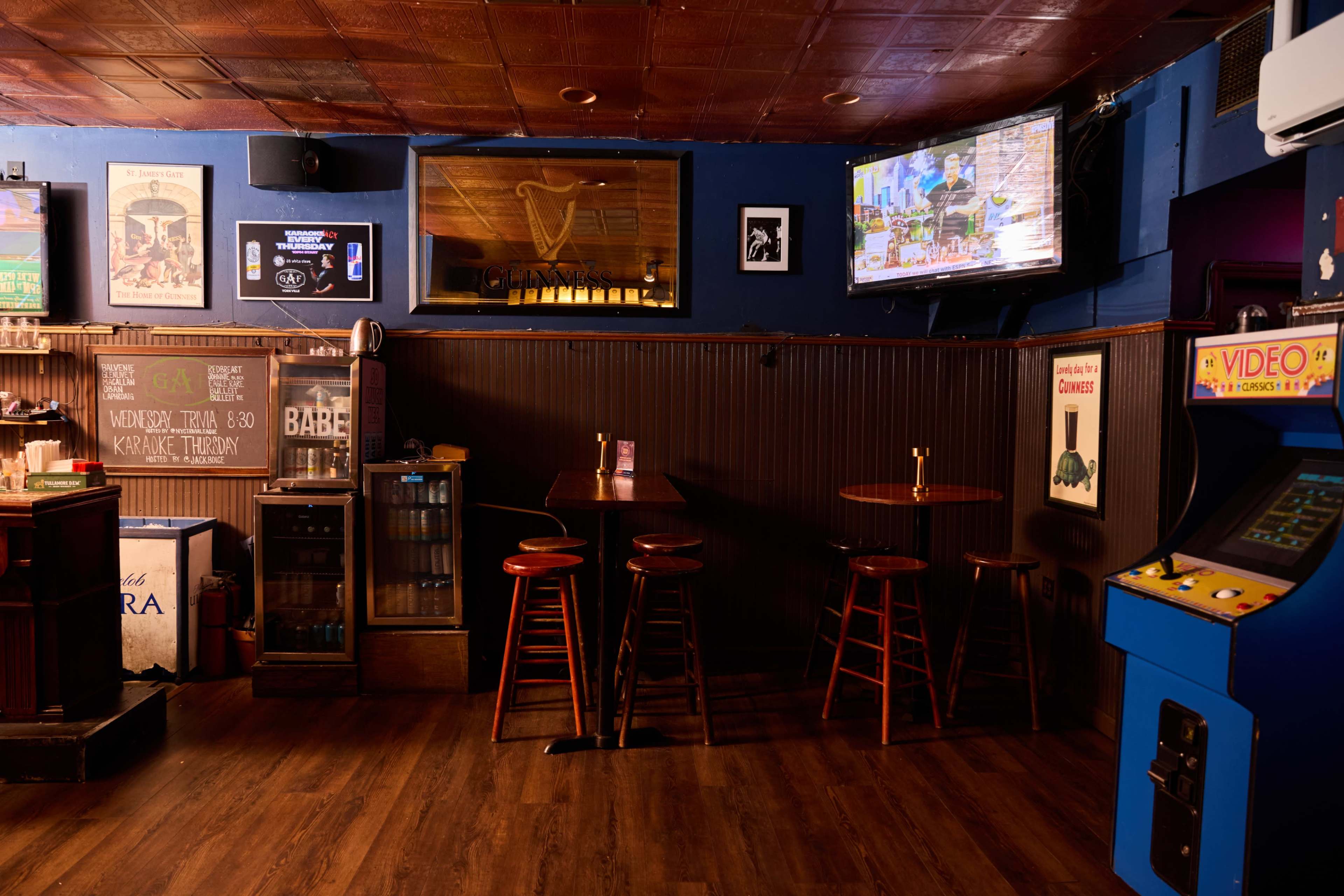The image shows the interior of a bar featuring a few wooden stools, a small round table, a television on the wall, and a video game machine in the corner.