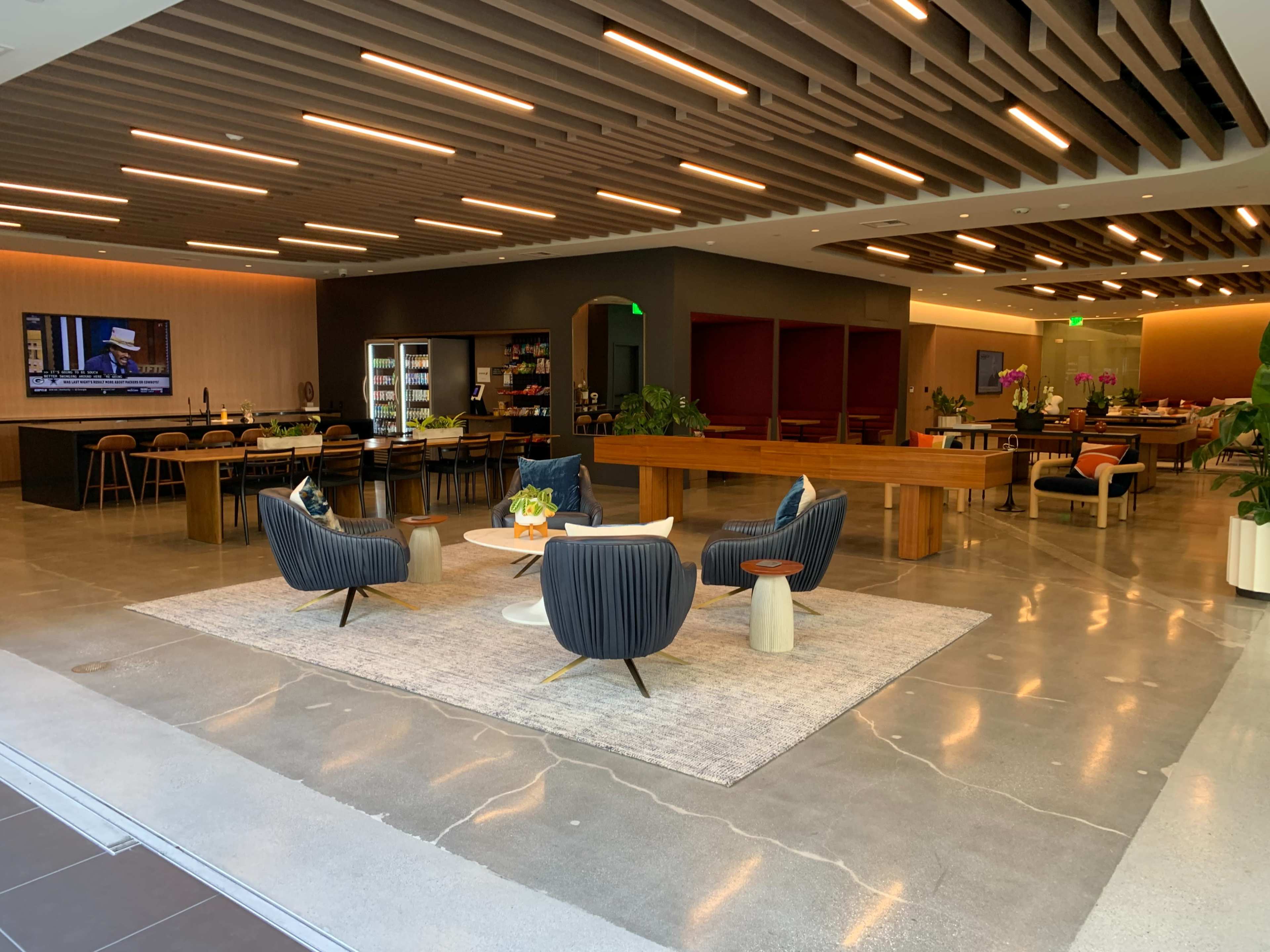 The image shows a modern office lounge area with stylish seating, a large communal table, a TV mounted on the wall, and plants for decoration.