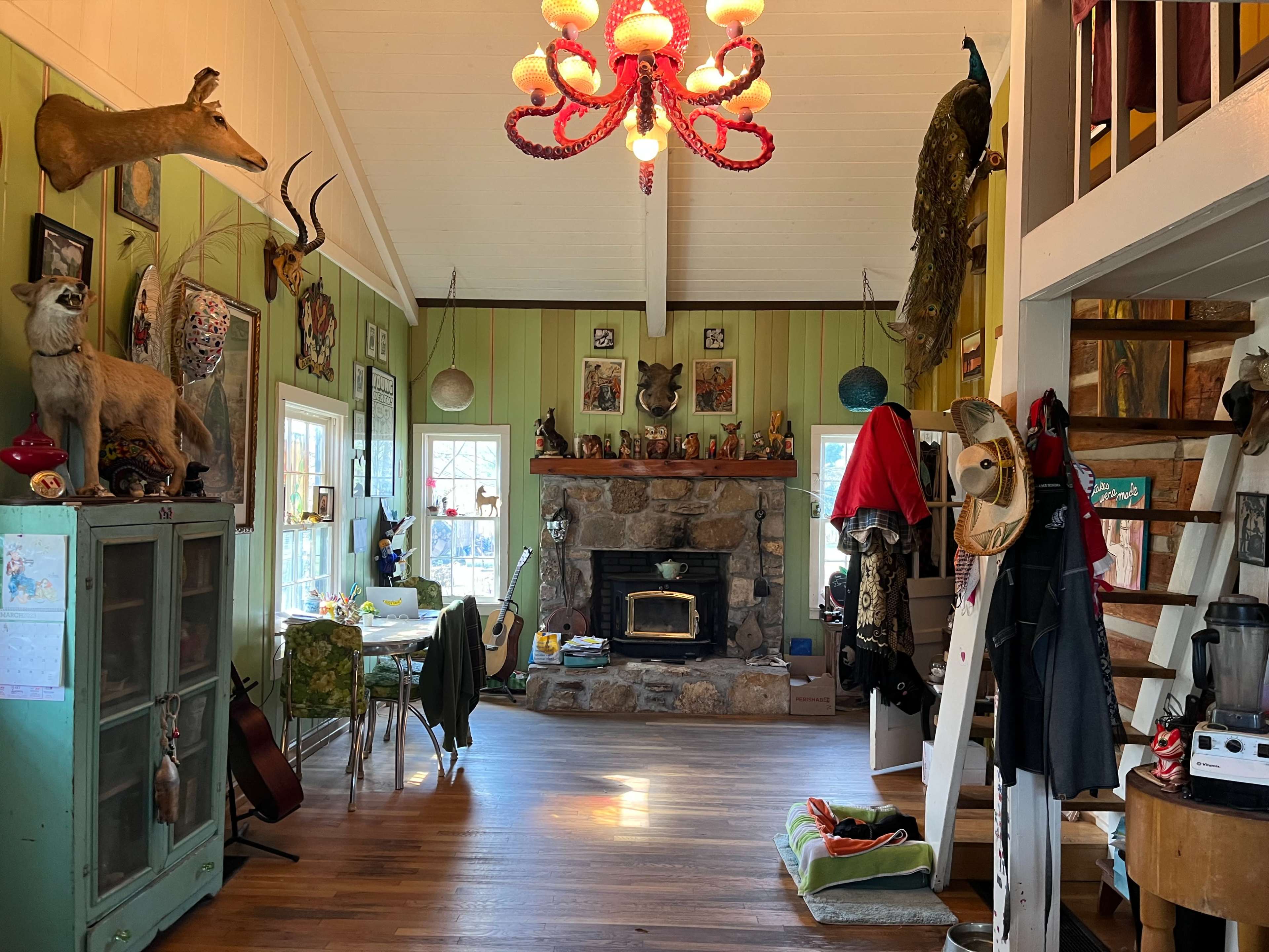 The interior of a cozy room features a stone fireplace, eclectic decorations, a colorful chandelier, and a mix of furniture and items scattered around.