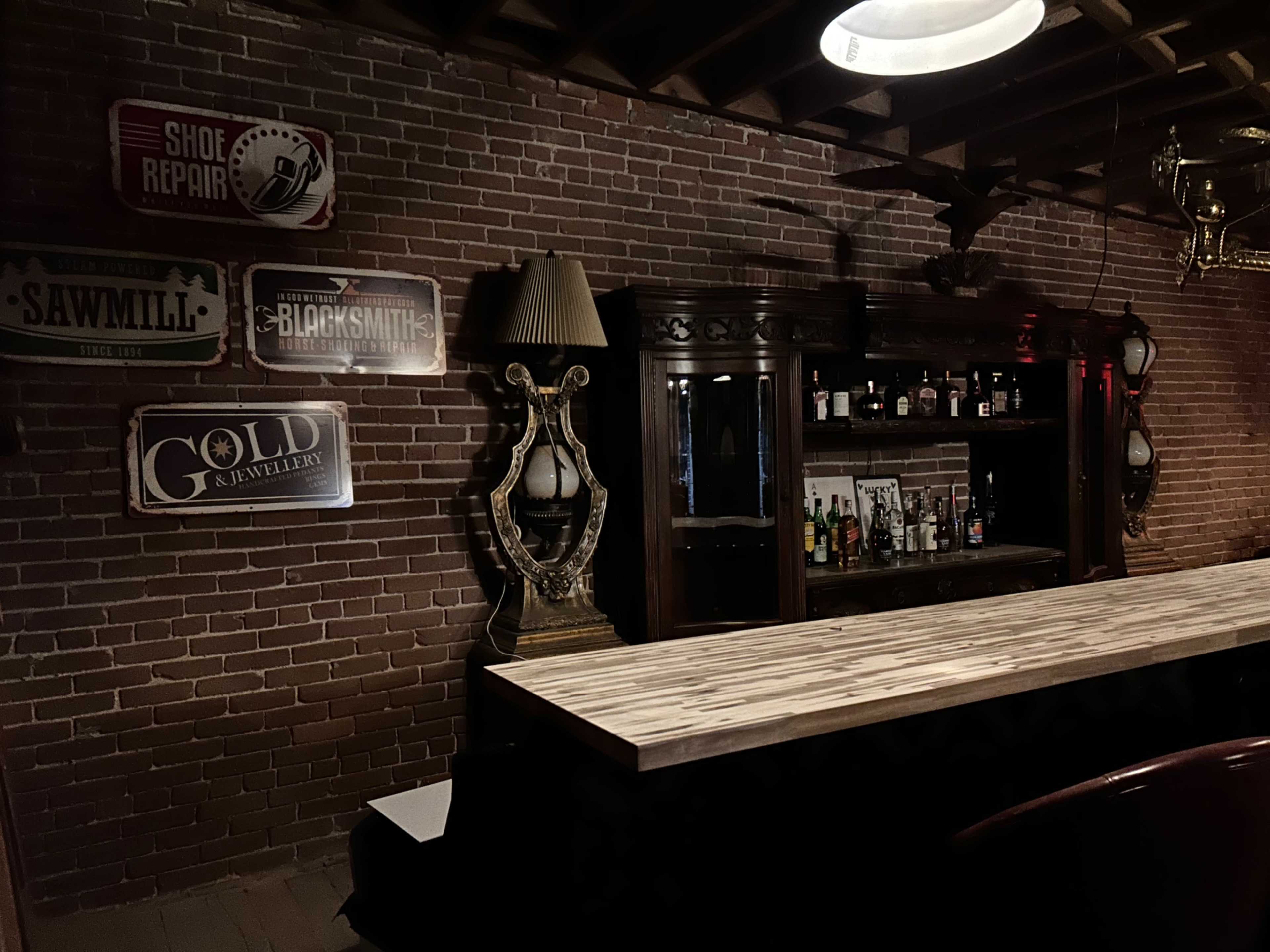 The image shows a dimly lit bar area with a wooden countertop, an antique cabinet filled with bottles, and vintage signs on the brick wall.