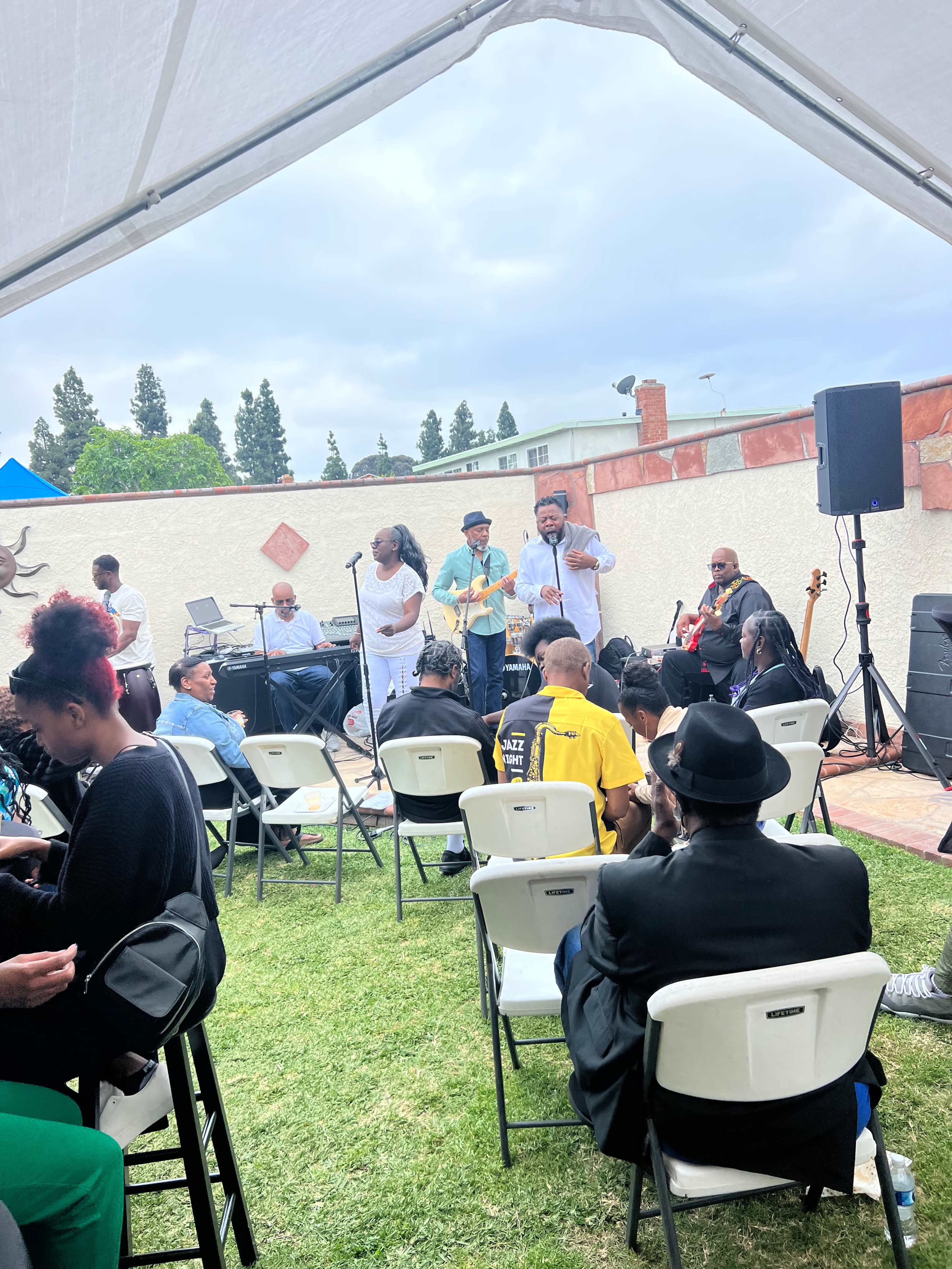 A group of musicians performs on a small stage while an audience sits in folding chairs under a tent.