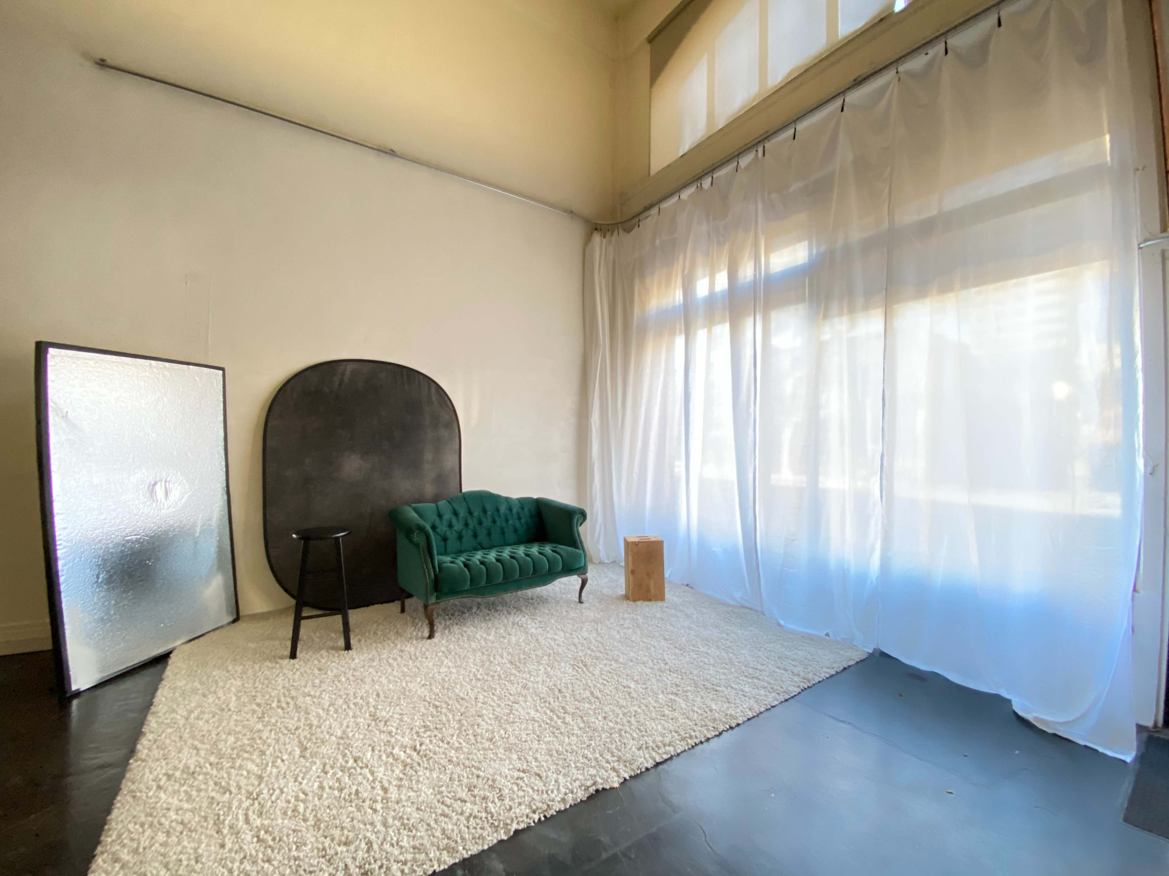 The scene features a bright studio with a green velvet sofa, a small black stool, a wooden cylinder, and sheer white curtains allowing natural light.
