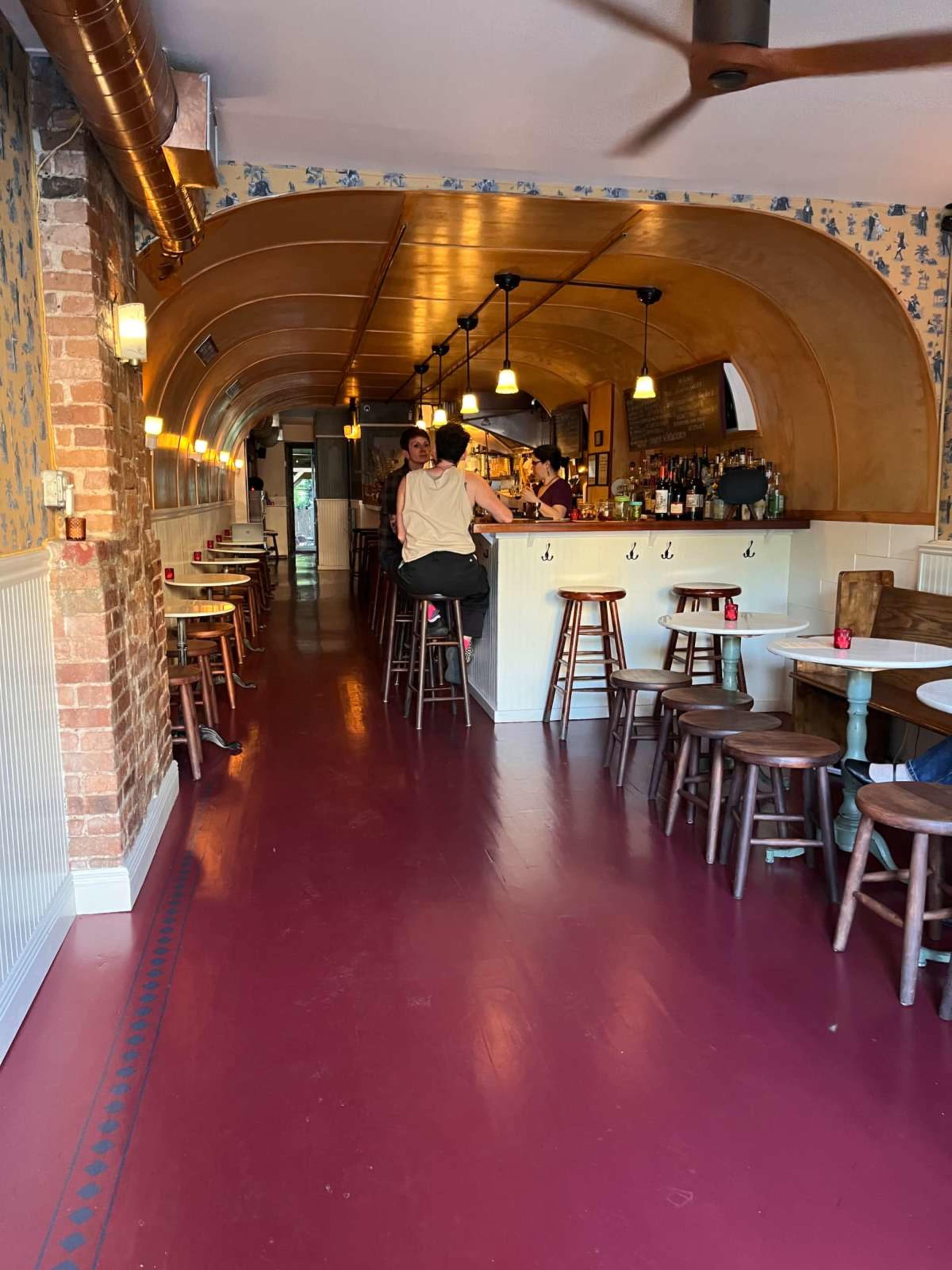 The image shows the interior of a restaurant with a long, narrow design featuring a bar at one end, wooden stools, and a variety of tables along the sides.