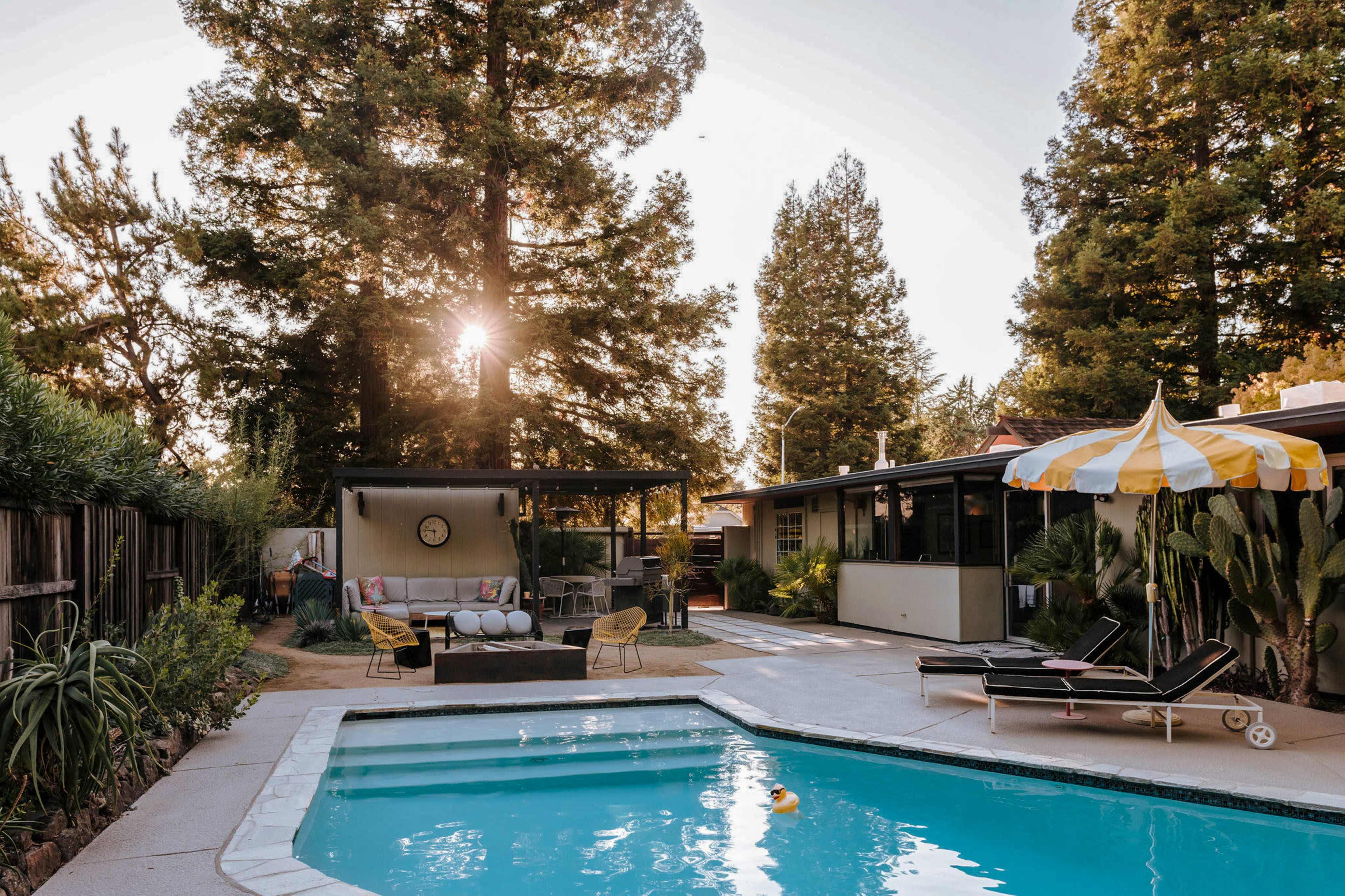 A backyard pool area is surrounded by trees, featuring a seating area, a sun umbrella, and a lounge chair beside the water.