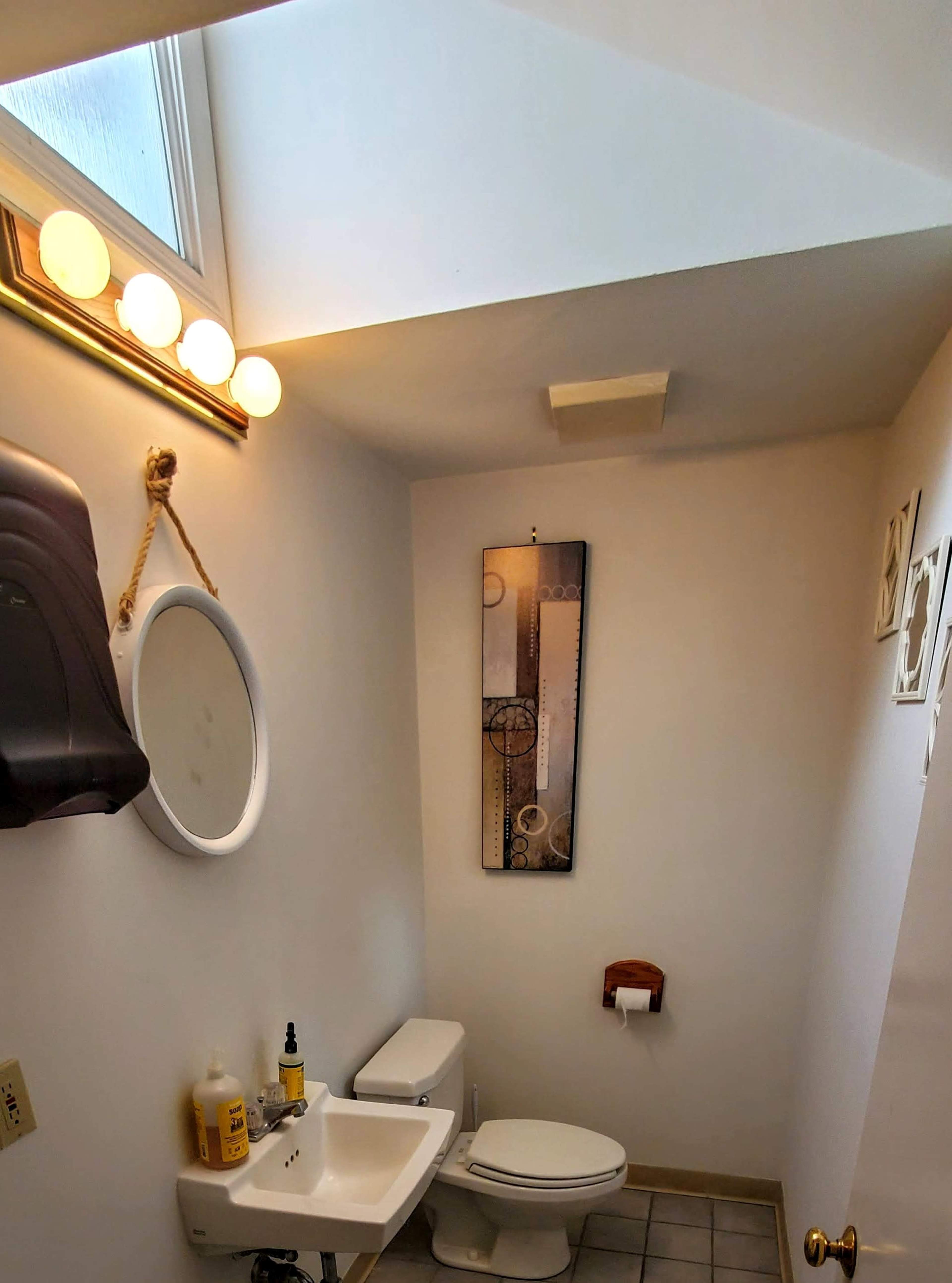 A small bathroom with a toilet, a sink, a round mirror, and a wall-mounted paper towel dispenser, illuminated by overhead lights.
