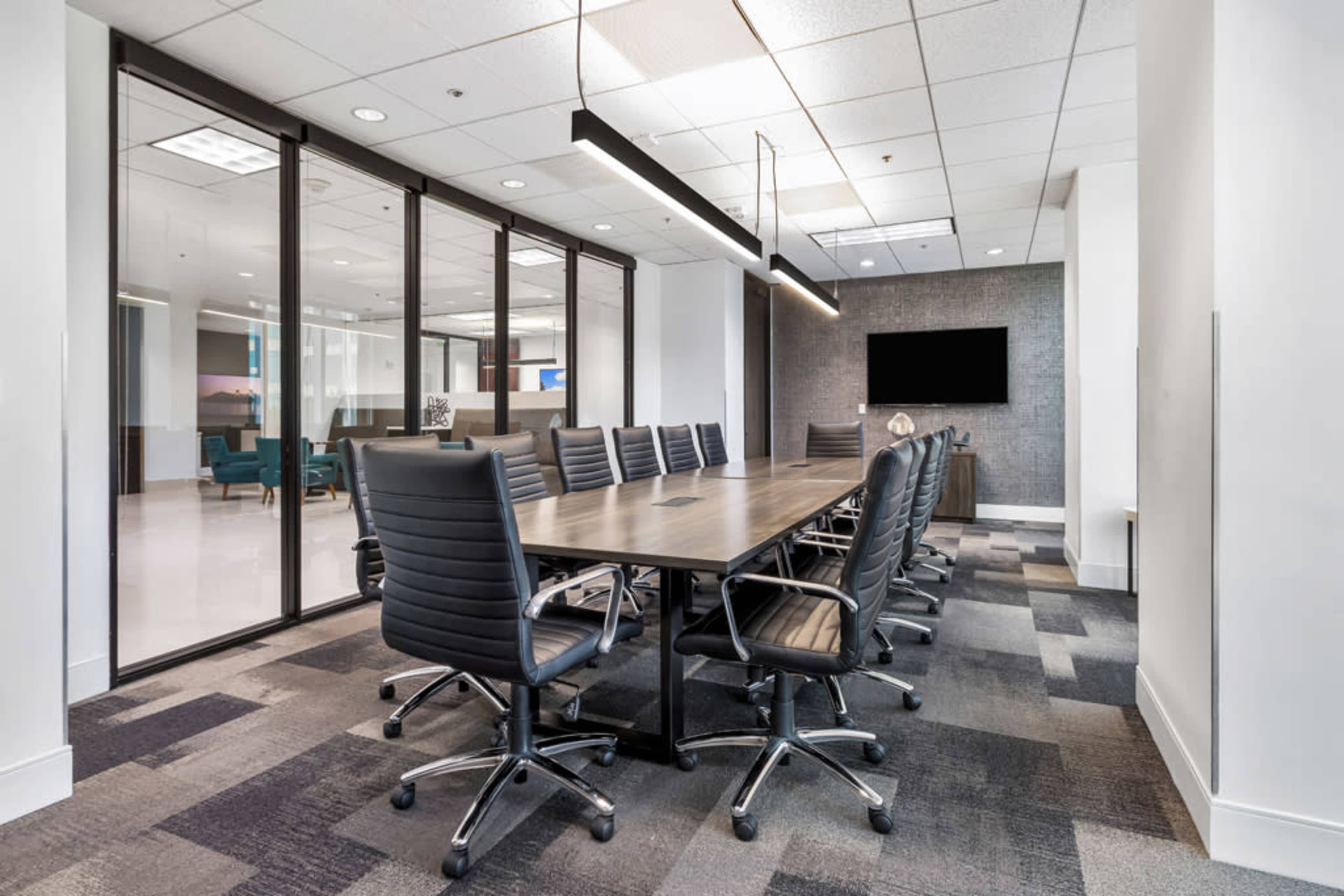 A large conference room features a long wooden table surrounded by black leather office chairs, with glass walls and a television mounted on the wall.