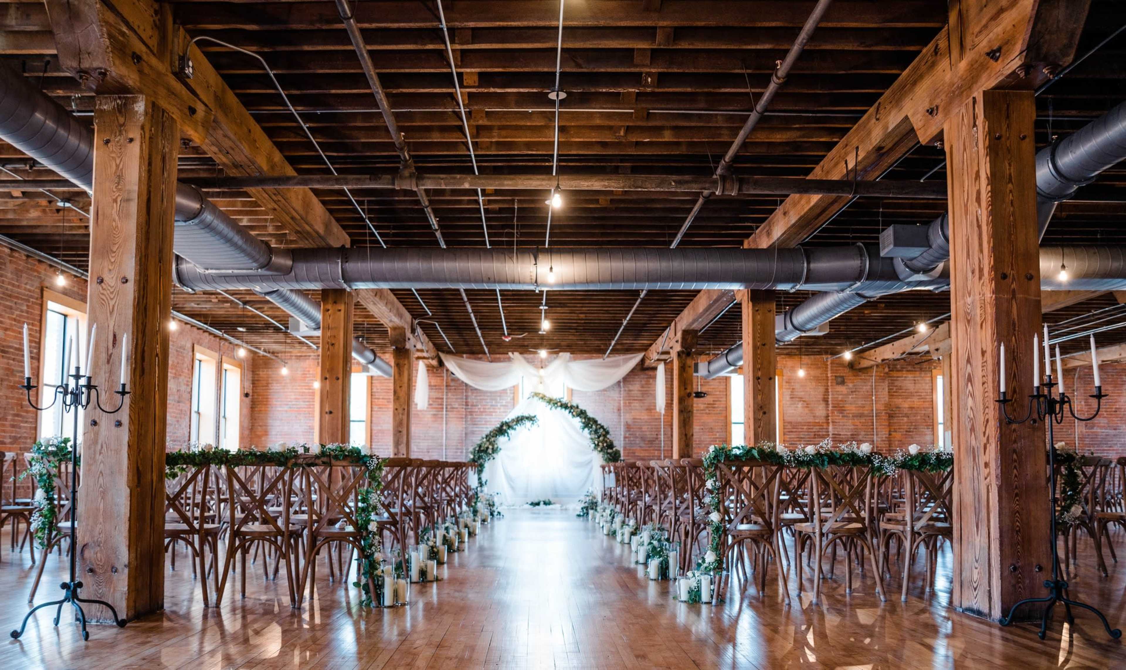 A spacious indoor wedding venue with wooden beams, arranged rows of chairs, and a floral arch at the front.