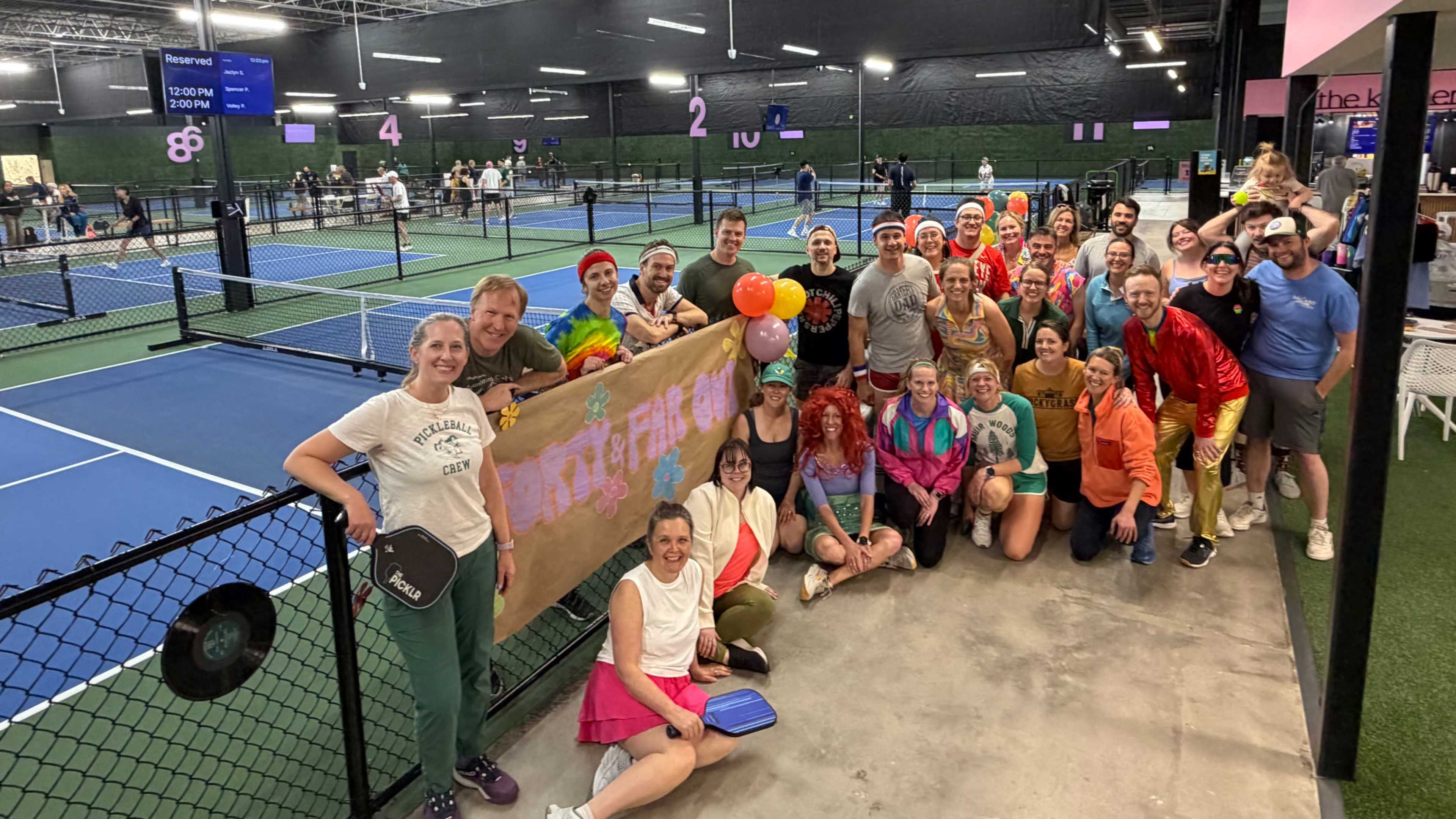 A large group of people dressed in colorful costumes poses together on a tennis court, with several courts visible in the background.
