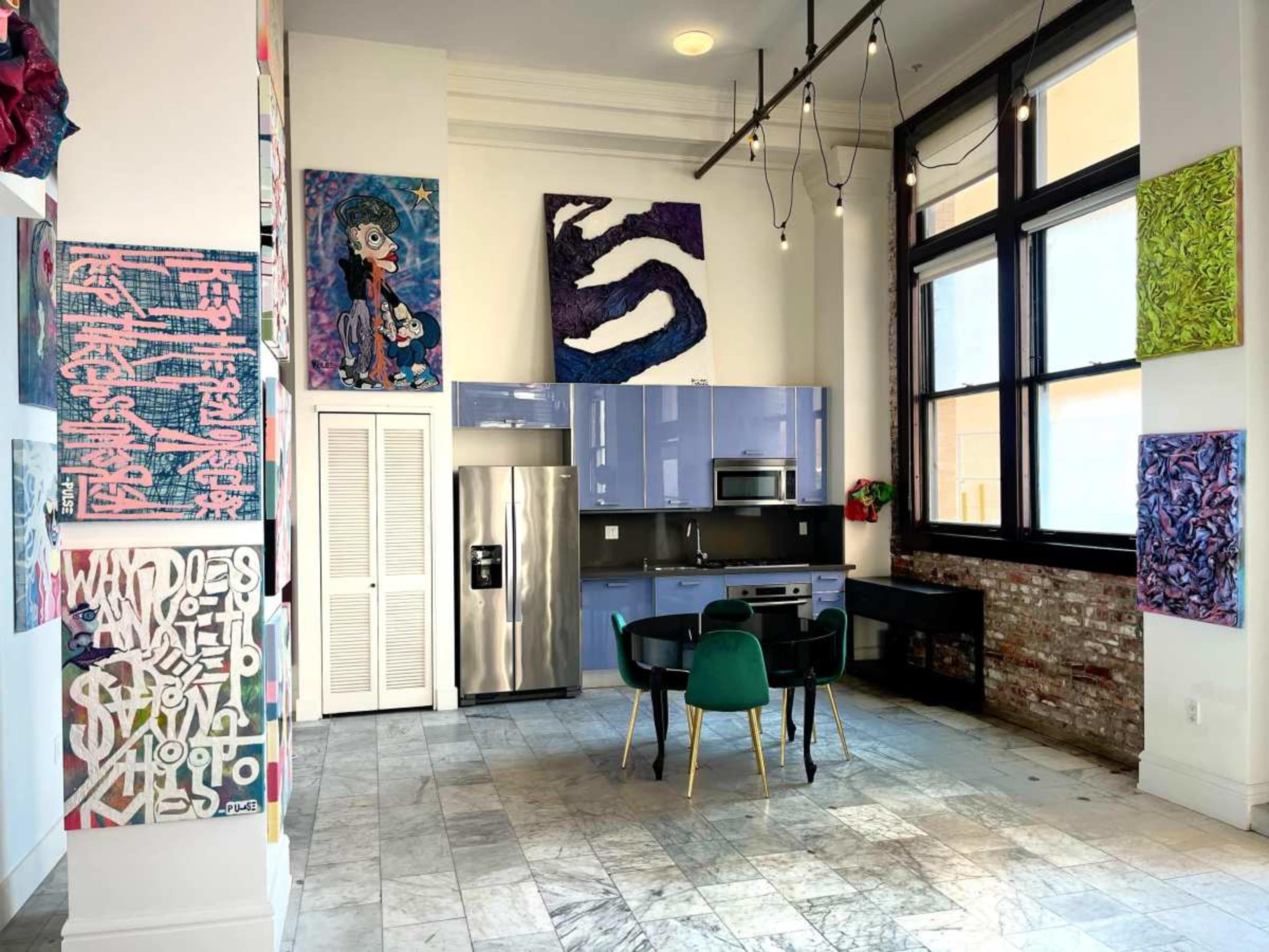 The image shows a modern kitchen and dining area with colorful artwork on the walls, featuring a refrigerator, microwave, and a dining table with green chairs.
