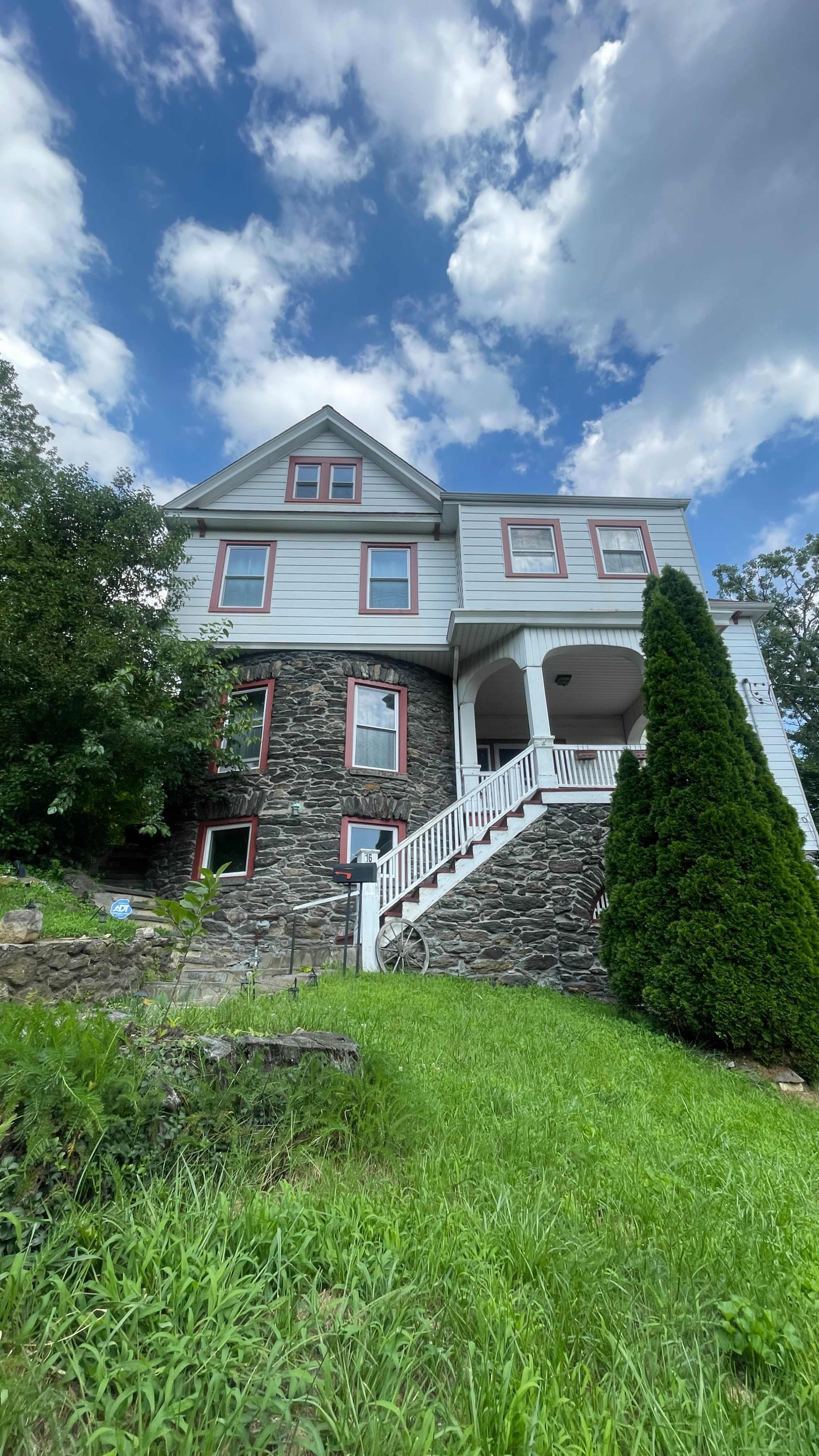 A large, two-story house with a stone and siding exterior is situated on a grassy hillside, featuring multiple windows and a front porch with steps.