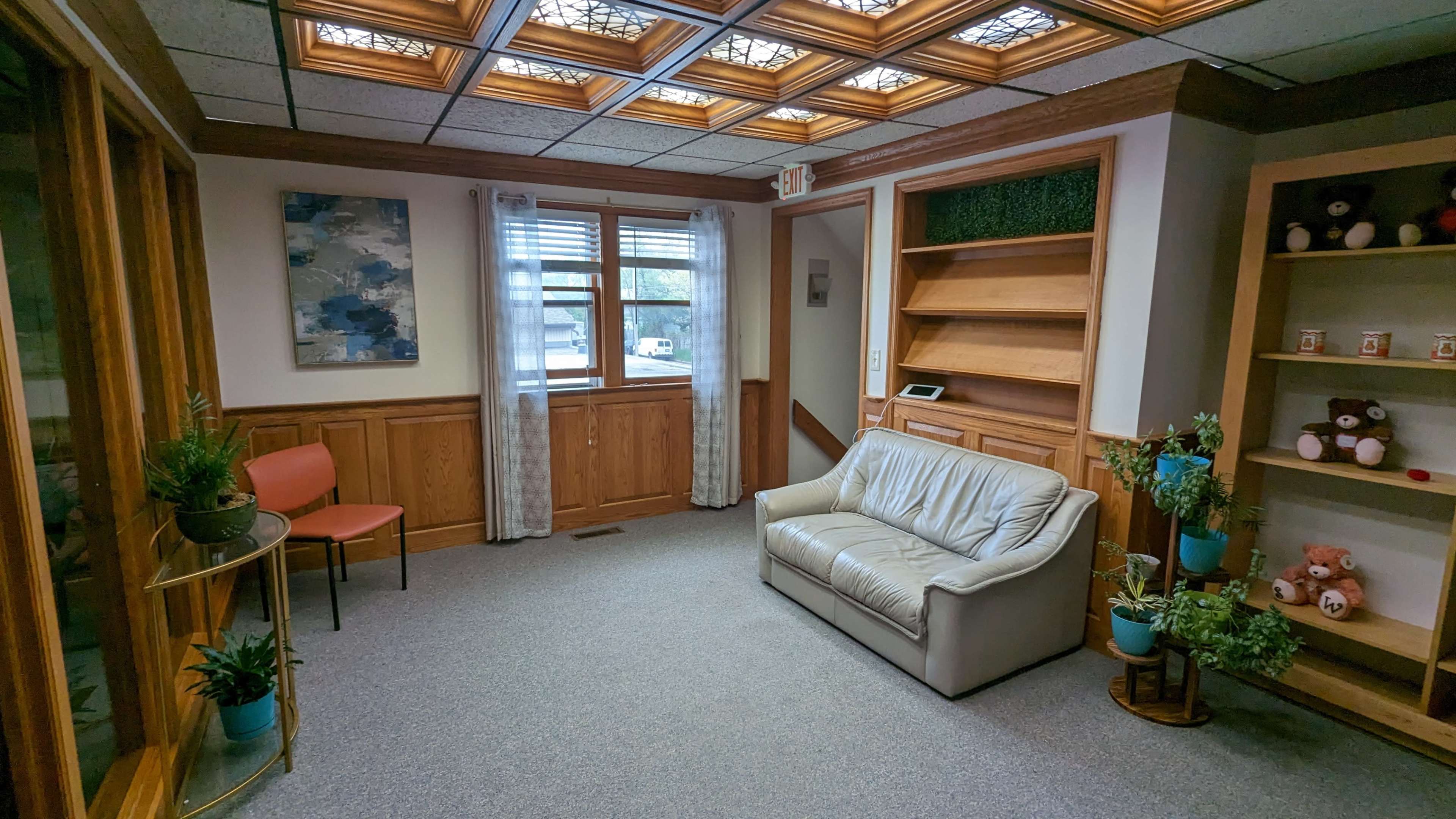 The image shows a well-lit waiting room with a beige leather sofa, a couple of chairs, decorative plants, and a bookshelf.