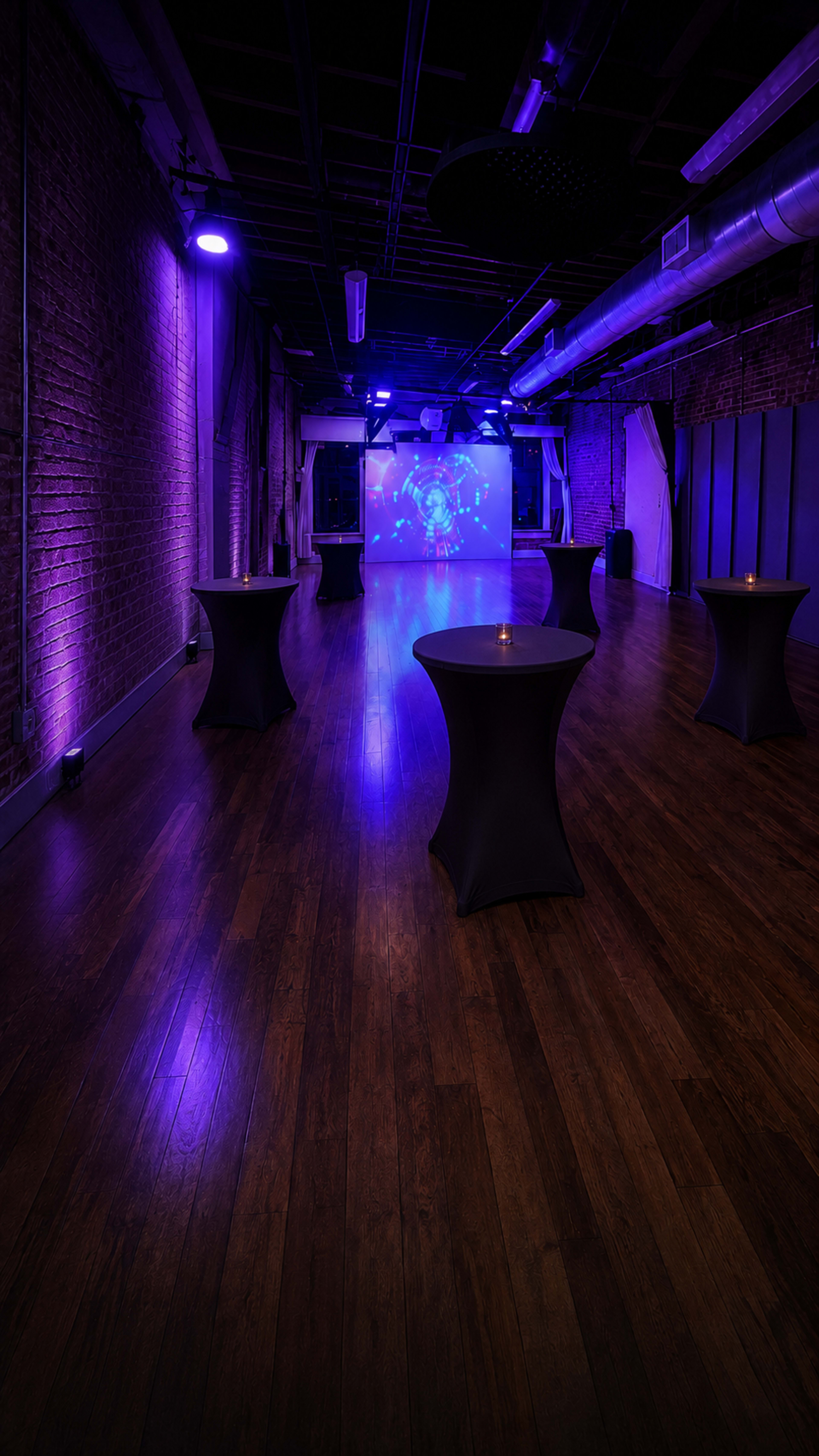 The image shows a spacious event venue with purple lighting, featuring several cocktail tables arranged around a projected screen at one end.