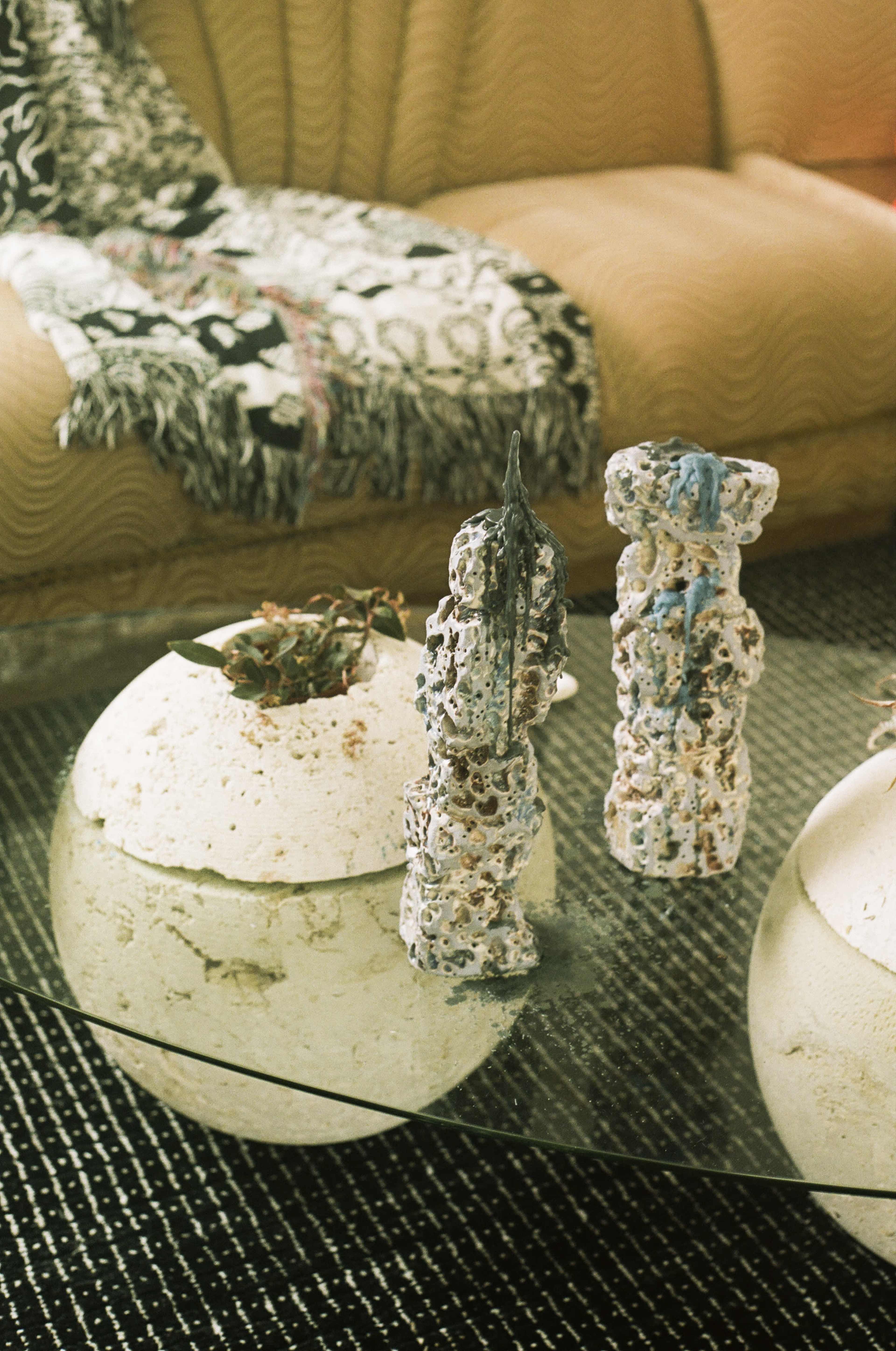The image features two textured ceramic sculptures on a glass table, accompanied by a potted plant and a patterned textile draped over a sofa in the background.