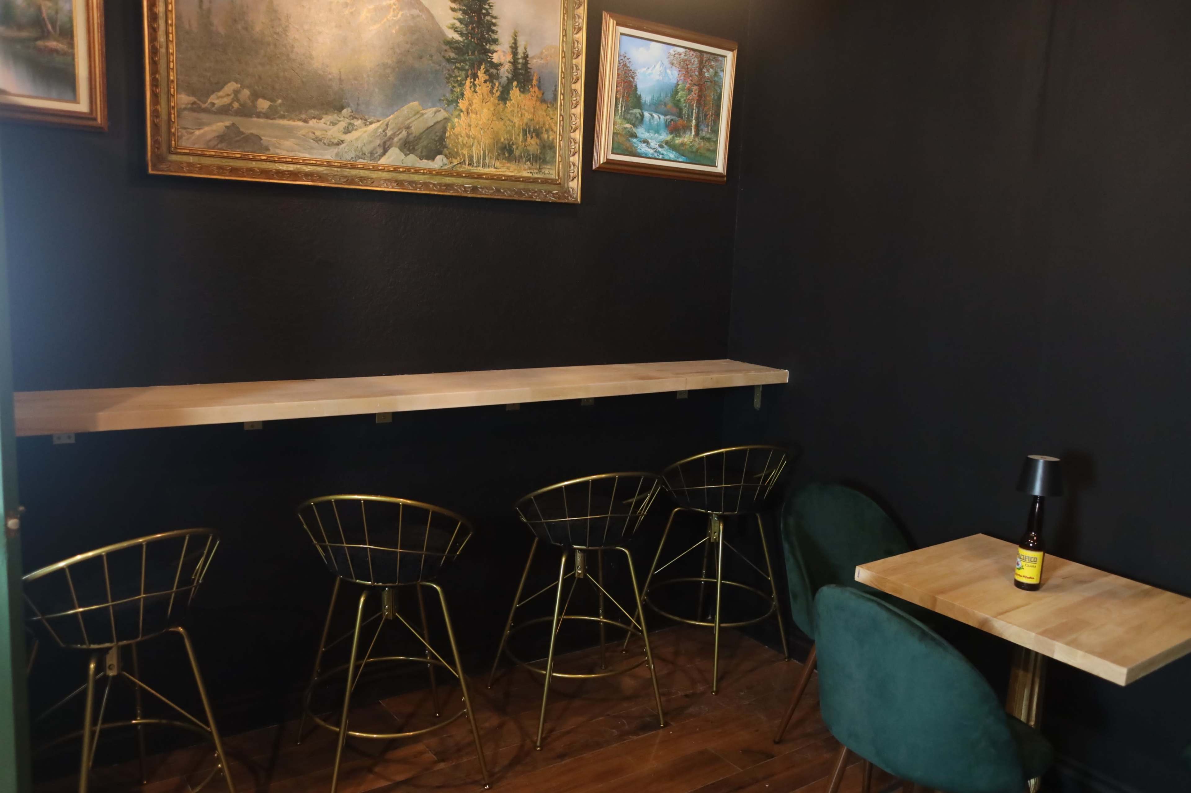 The image shows a small cafe space with a wooden table and four gold wire bar stools along a wall adorned with two framed landscape paintings.