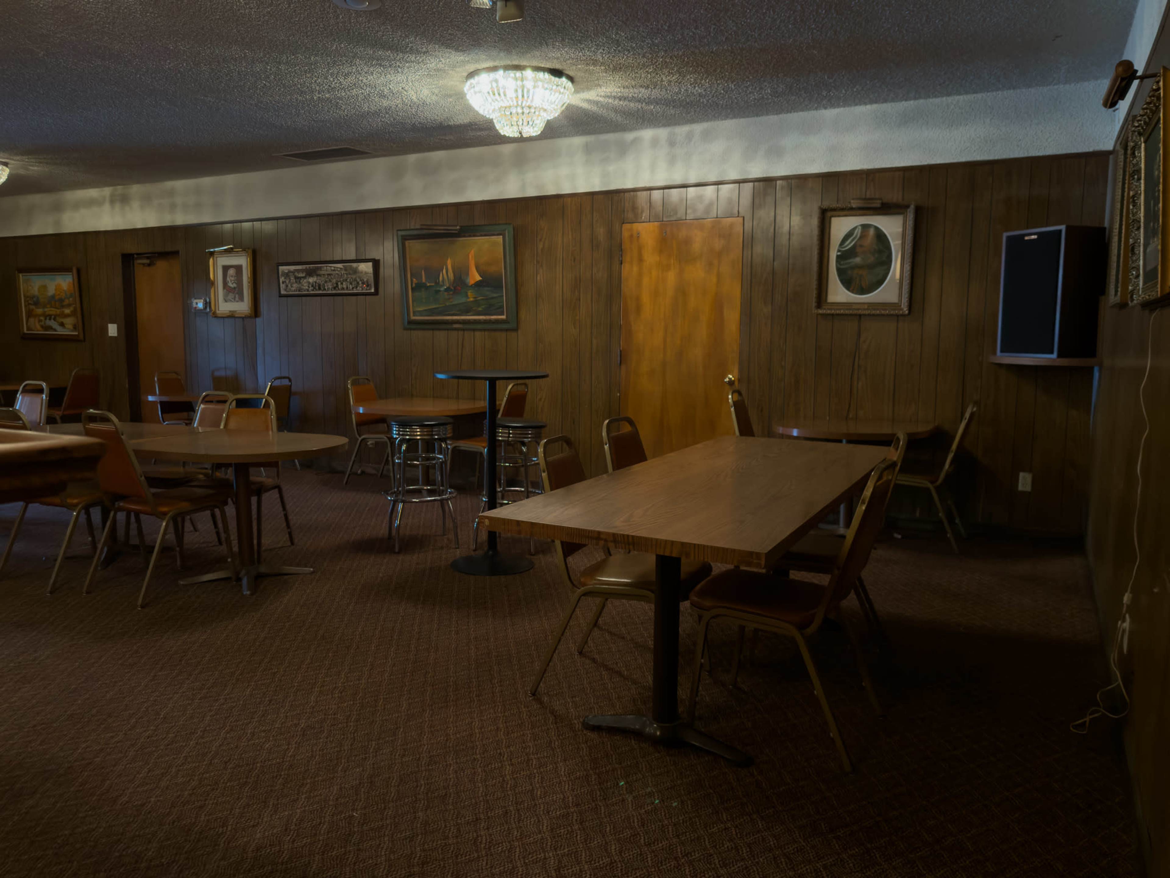 The image shows a sparsely furnished room with several tables and chairs, wooden paneling on the walls, and dim lighting from a chandelier.