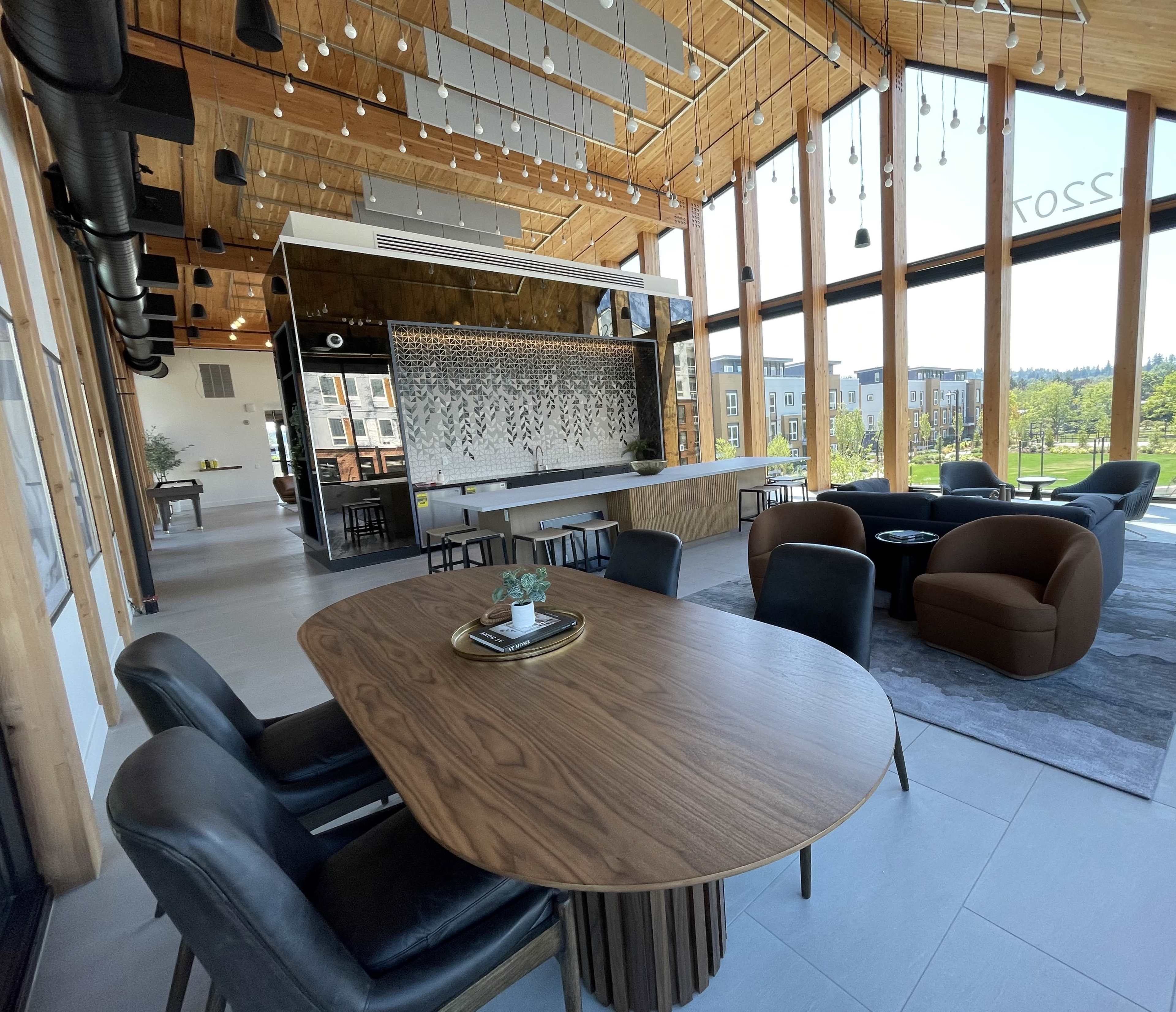 A modern indoor space featuring a large wooden dining table, black chairs, and a lounge area with a round sofa, all set against expansive windows revealing a view of the outdoors.