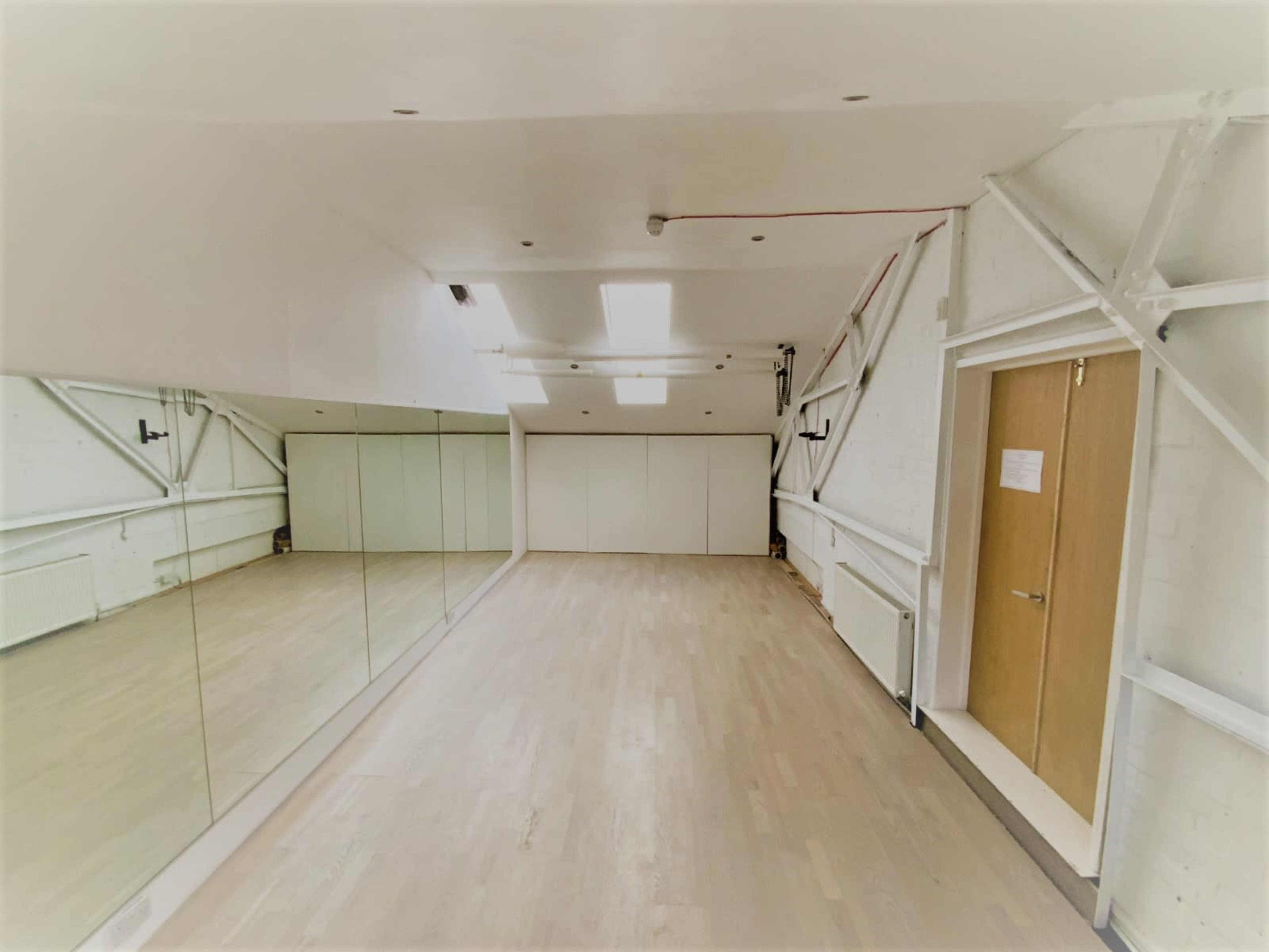 A spacious, well-lit room with a large mirror along one wall and a door on the right, featuring white walls and wooden flooring.